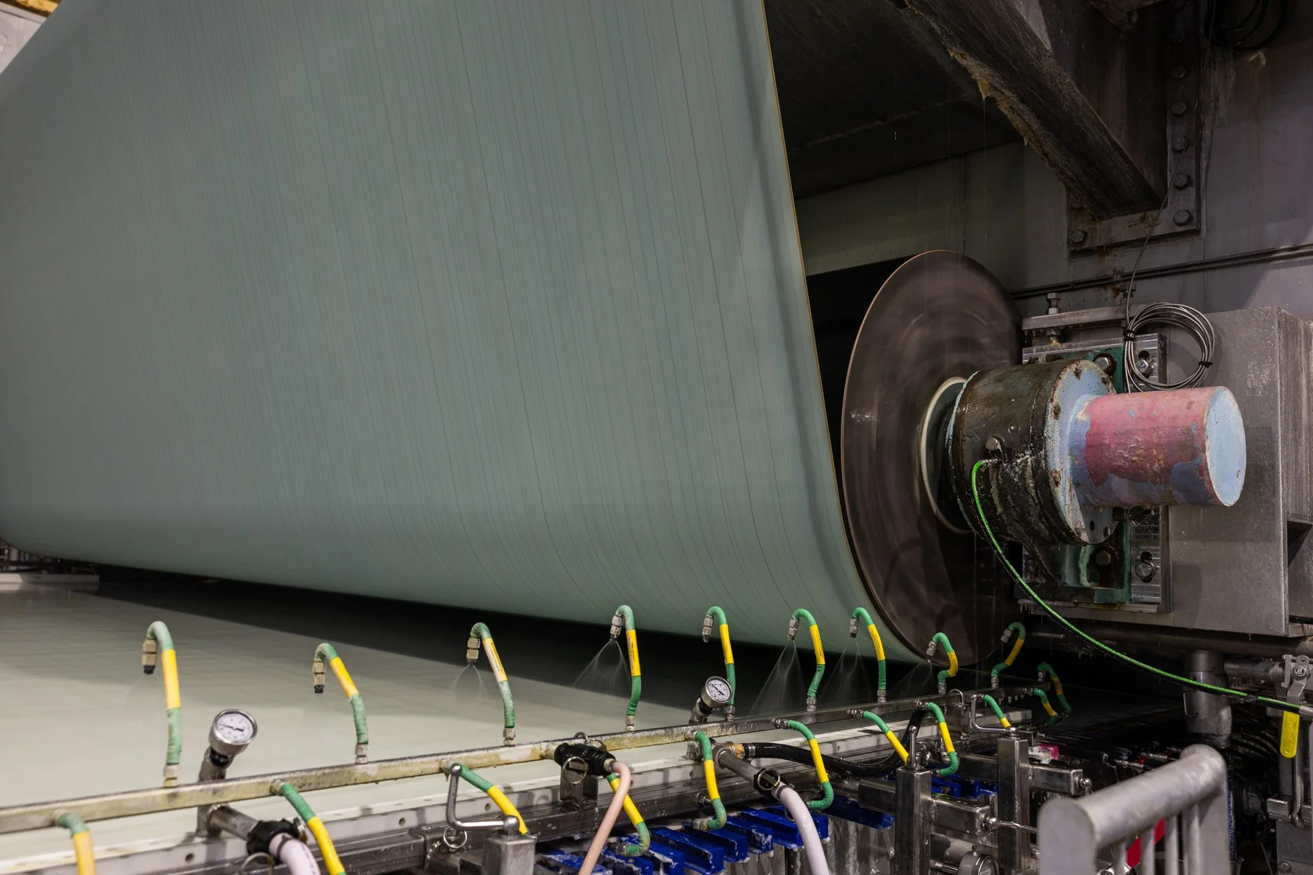 Industrial manufacturing process with a large cylindrical roller, pipes, gauges, and hoses in a factory setting.