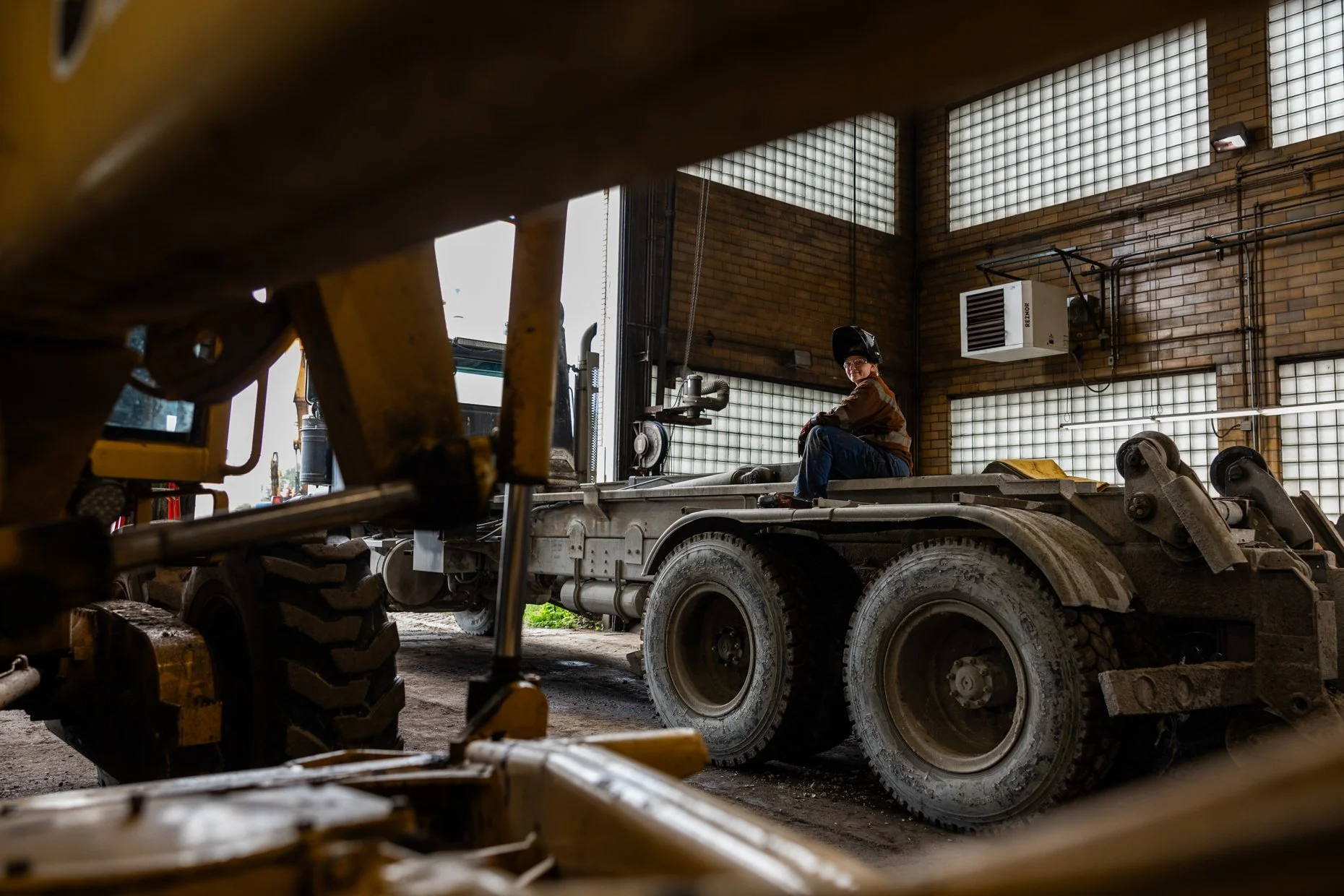 A worker wearing safety gear sitting on a flatbed truck inside an industrial warehouse with brick walls and glass block windows.