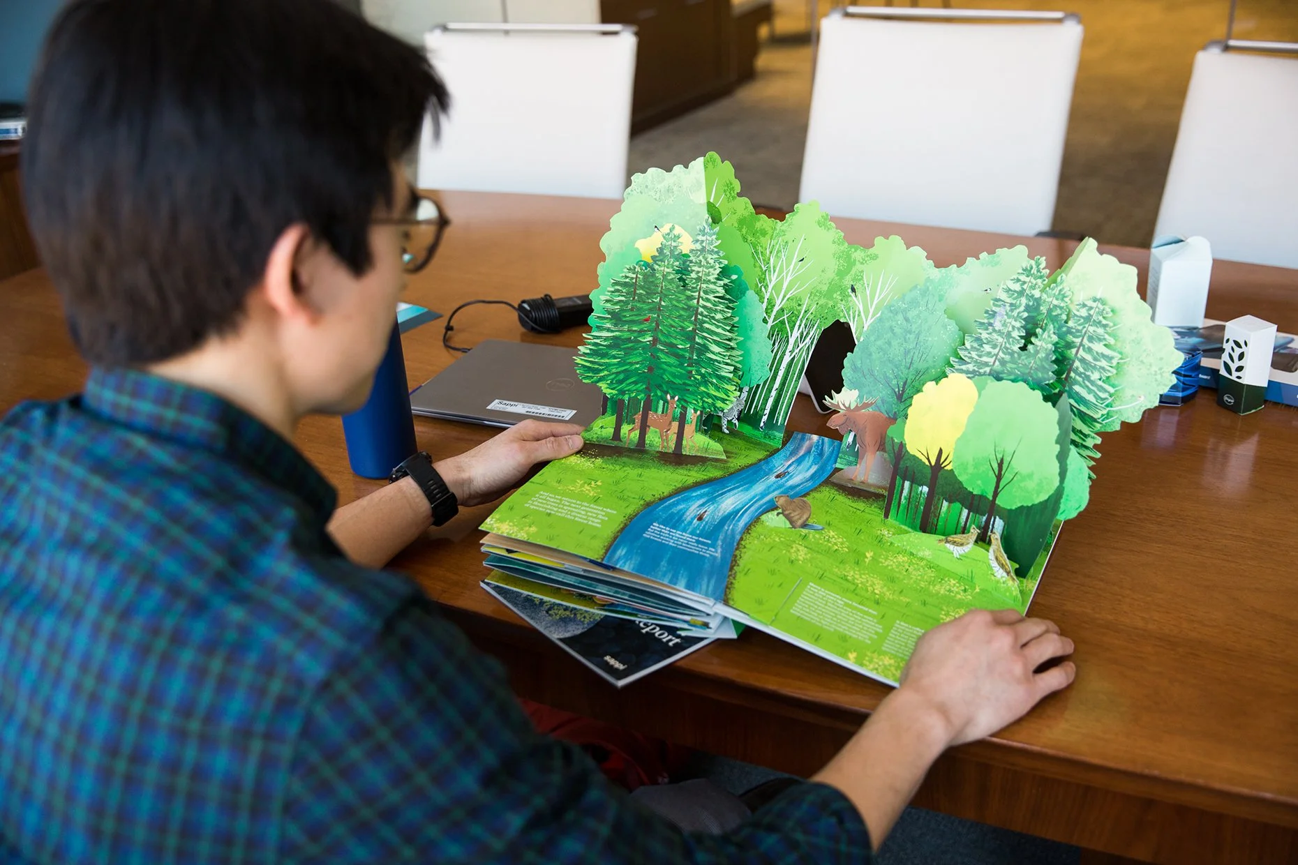 Person with glasses looking at a pop-up book featuring trees, a river, and forest animals on a wooden table in a conference room.