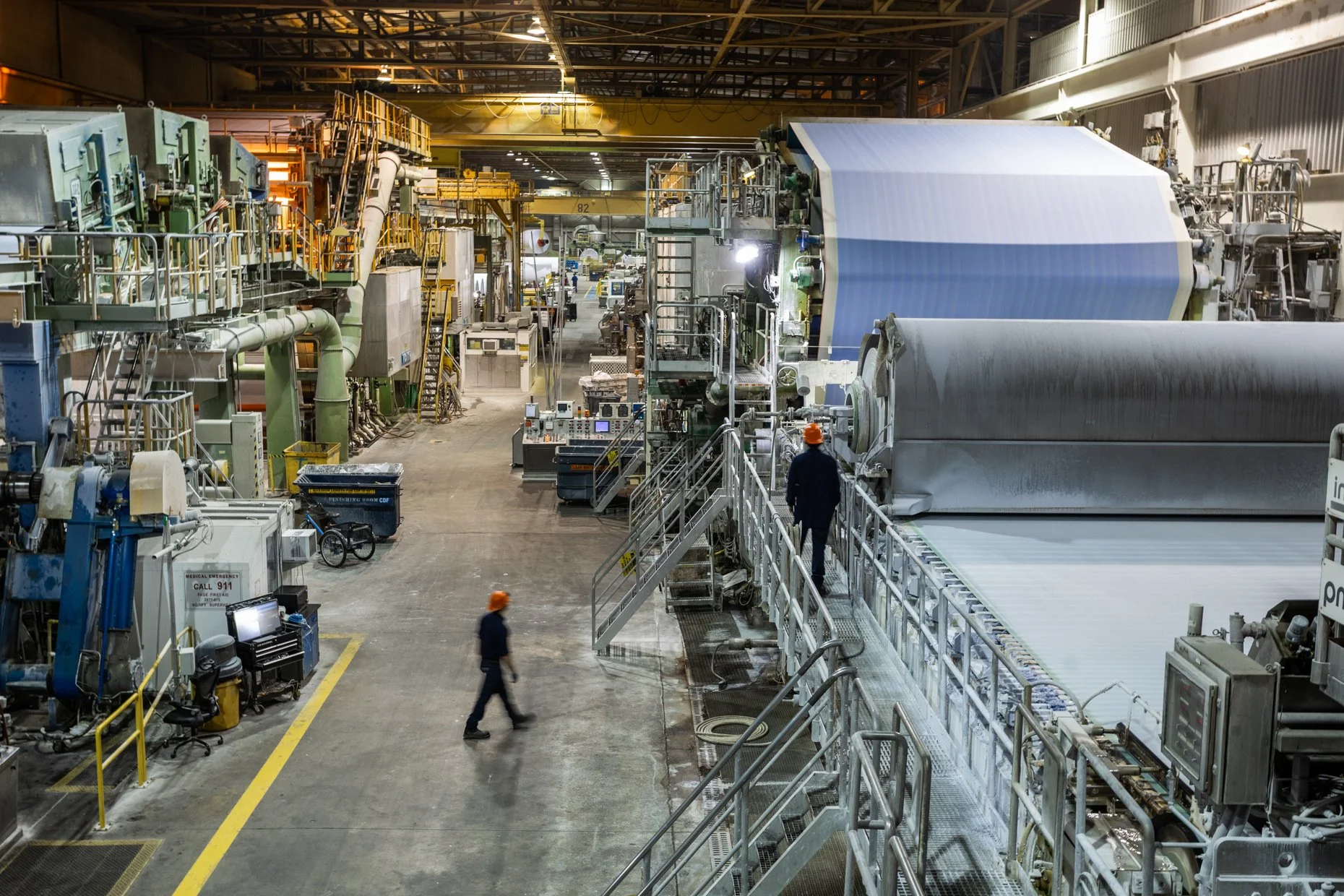 Inside an industrial factory with machinery for manufacturing large paper rolls, workers in orange safety helmets walking around, yellow safety railings, and overhead cranes.