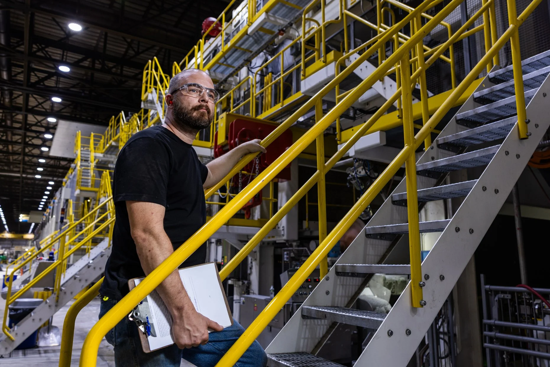 A man with safety glasses and a beard holding a clipboard, standing beside industrial machinery with yellow safety railings and stairs, in a warehouse or manufacturing facility.