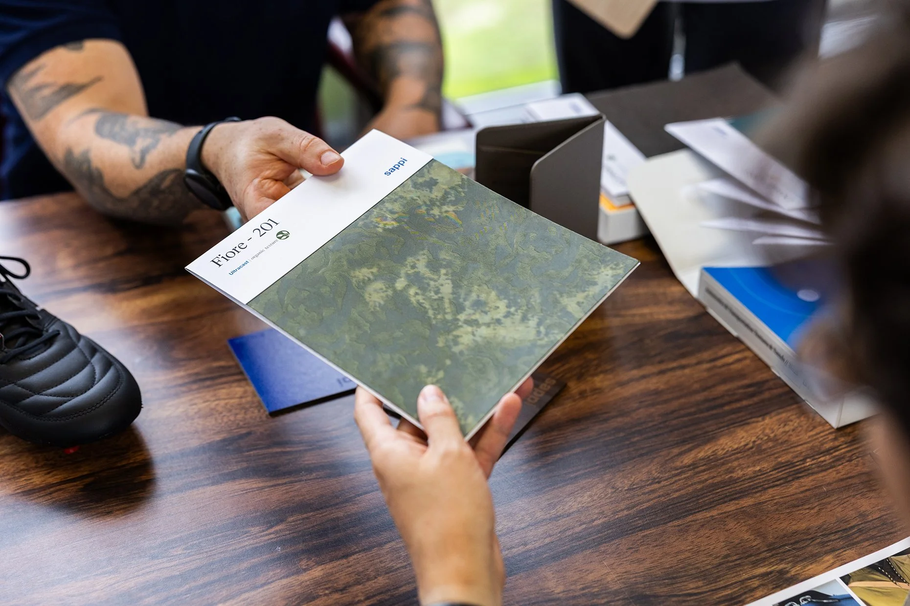Person holding a booklet with a forest image and the title 'Fiore - 201' at a table with papers and a shoe nearby.