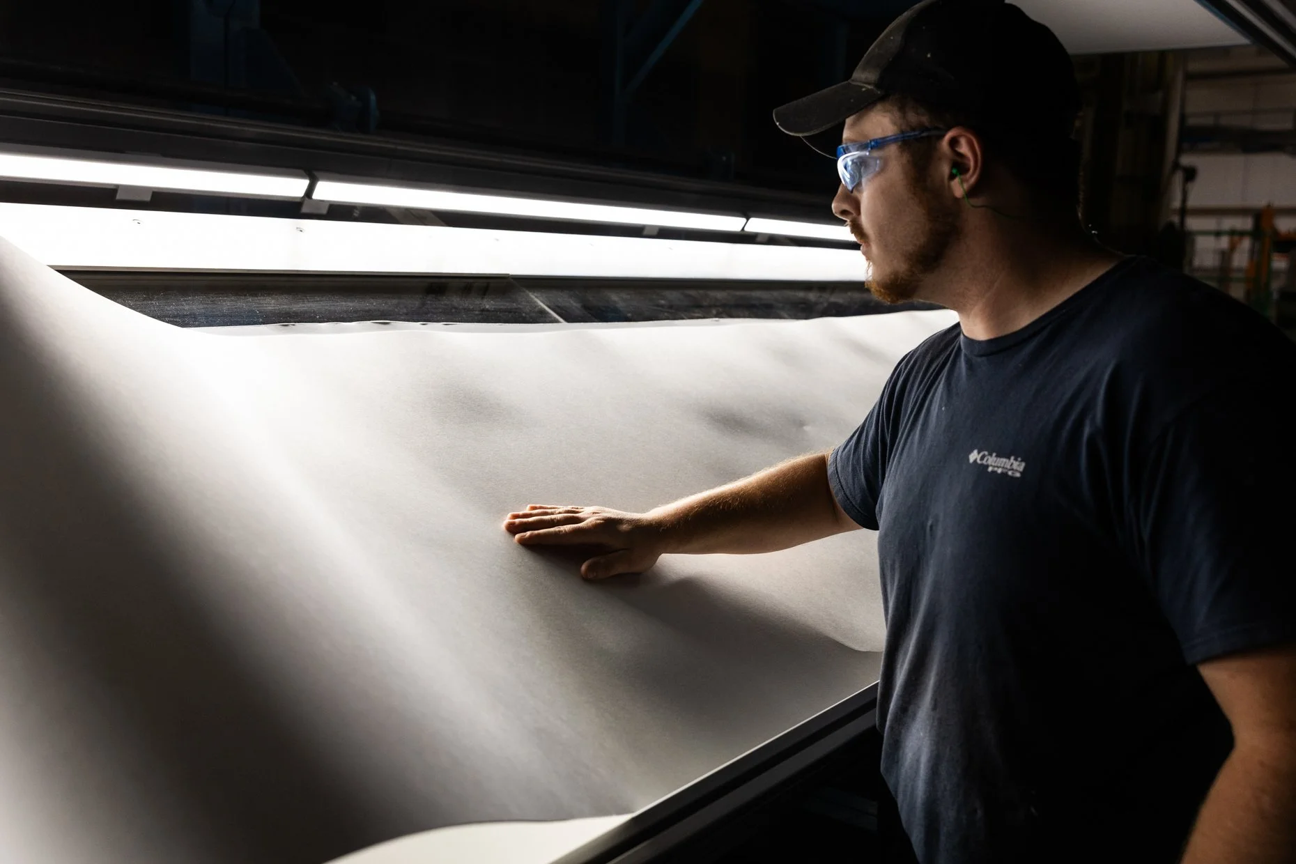 A man wearing safety glasses and ear protection is inspecting a large sheet of white fabric or paper on a long flat surface inside a factory or workshop.