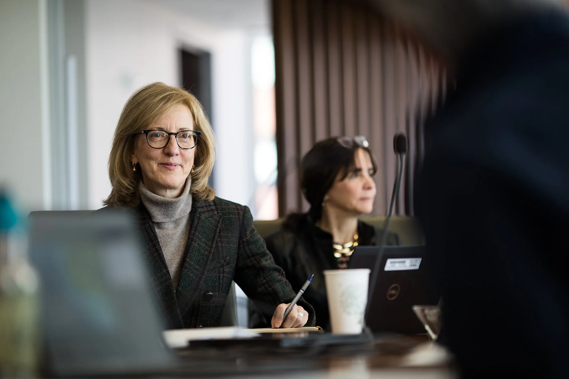 Two women seated at a conference table in a meeting room with wood paneling. The woman in the foreground has shoulder-length blonde hair, glasses, and is wearing a plaid blazer with a turtleneck. She is holding a pen and smiling slightly. The woman i