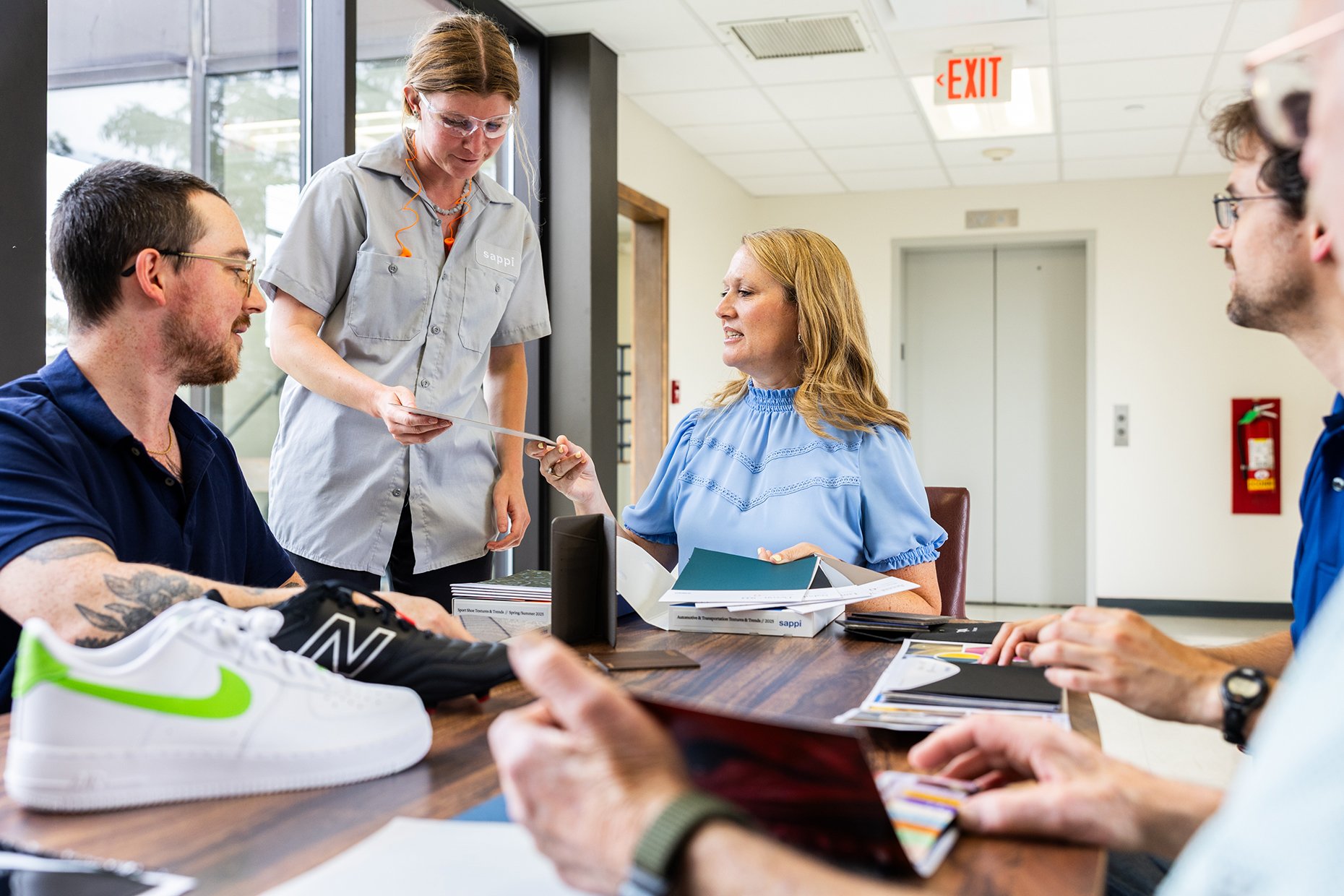 Group of five people sitting around a table in a meeting room, with a woman handing a document to a woman in a blue top. Sneakers placed in the foreground, and a fire extinguisher and exit sign visible in the background.
