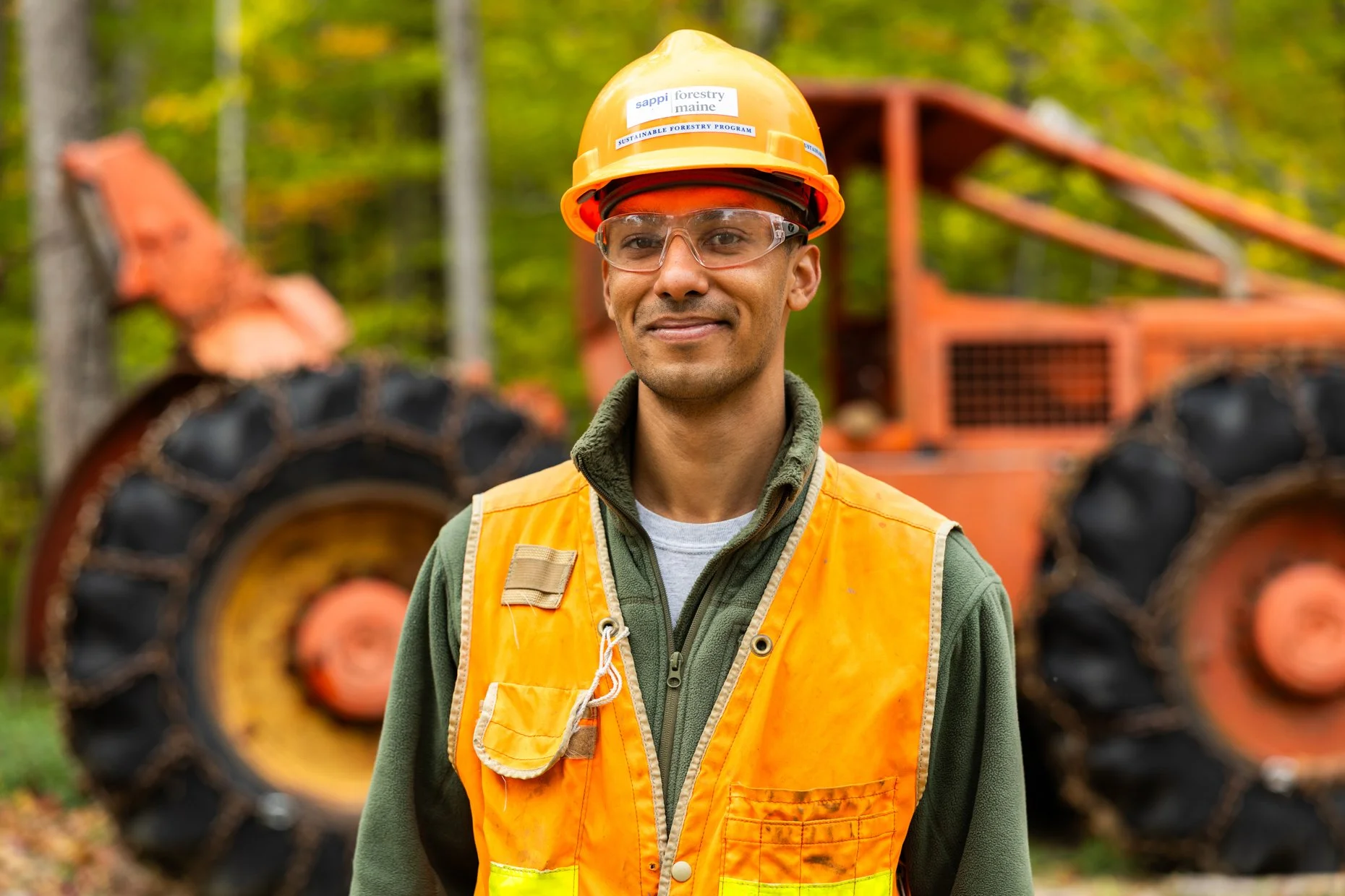 A man wearing safety glasses, an orange hard hat, and a yellow safety vest standing outdoors with a large orange forestry vehicle in the background.