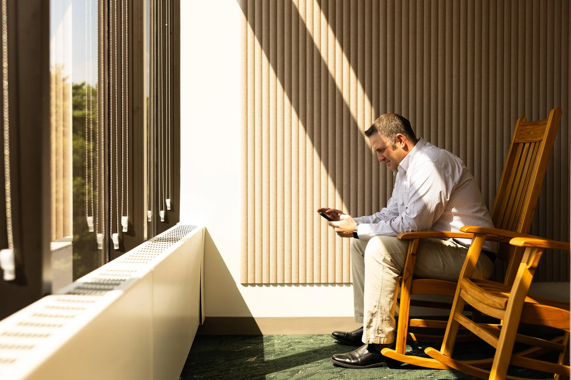 A man sitting on a wooden chair near a large window, looking at his smartphone, with sunlight casting shadows on a beige textured wall.