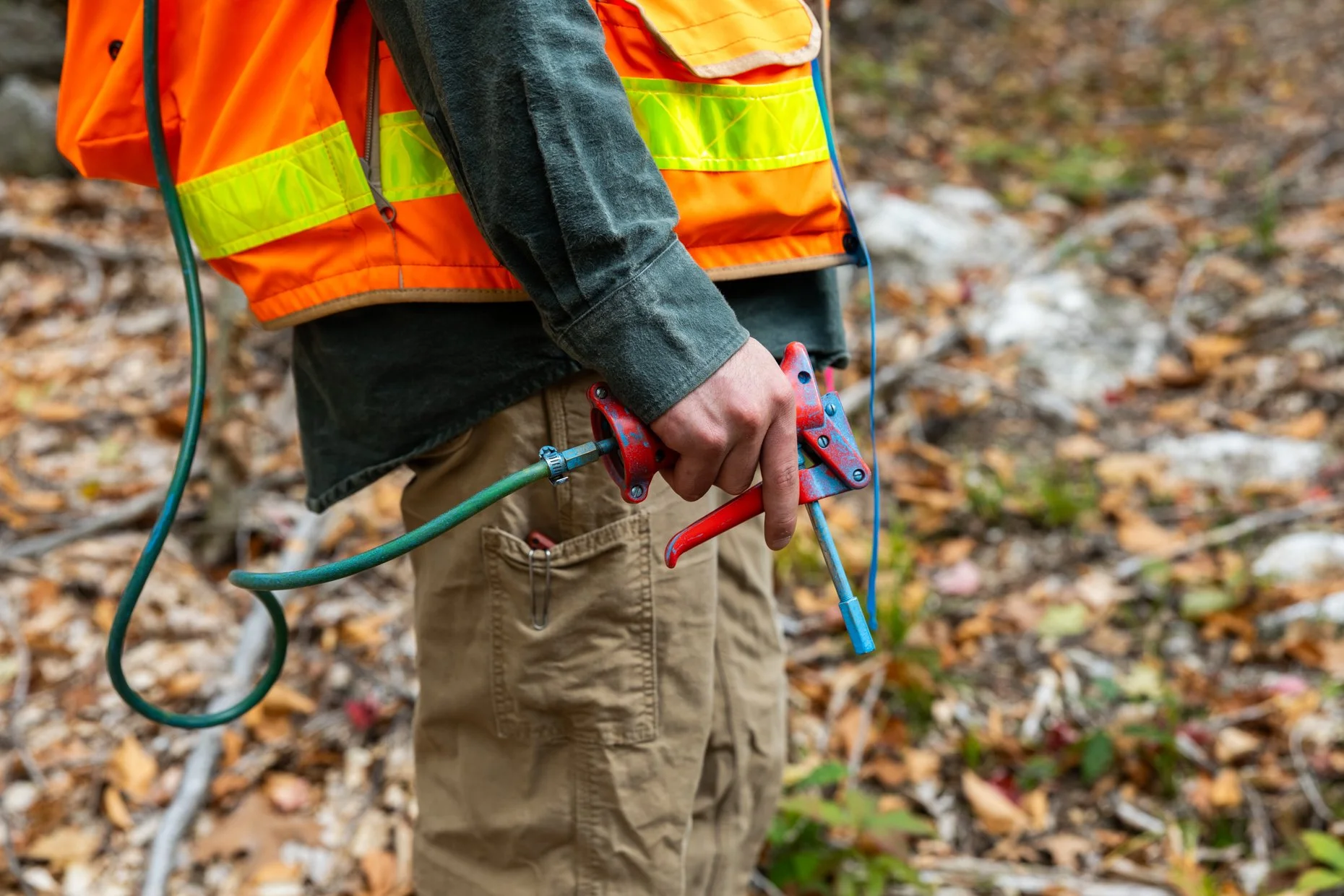 Person wearing an orange safety vest and khaki pants using a wrench and other tools outdoors among fallen leaves.