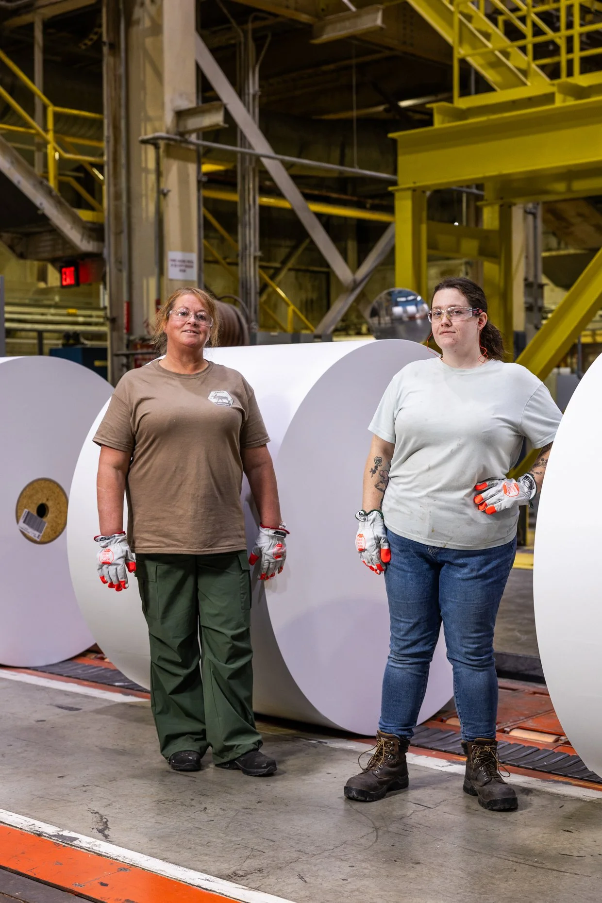 Two women in work uniforms and safety gloves standing in an industrial factory. Large rolls of material are behind them, and yellow industrial machinery is above.
