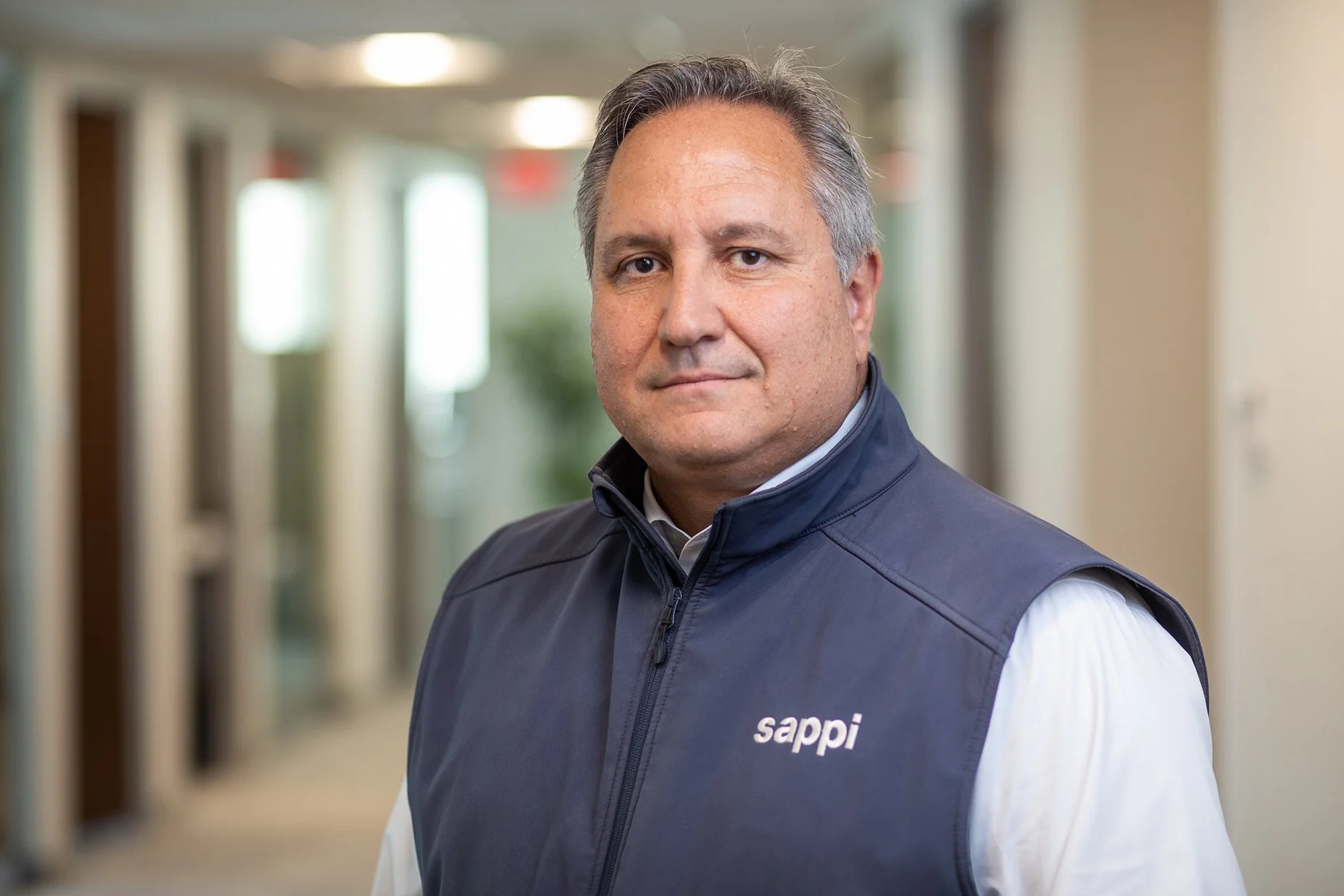 A man with short gray hair wearing a navy vest with the 'sappi' logo over a white shirt, standing in a brightly lit indoor hallway with blurred background.