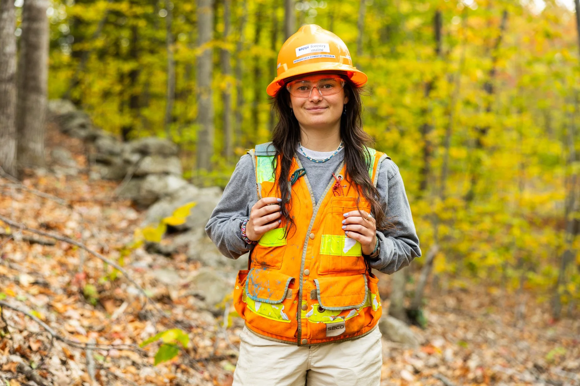 A woman wearing an orange safety vest, a yellow hard hat, and safety glasses standing in a forest with fallen leaves.