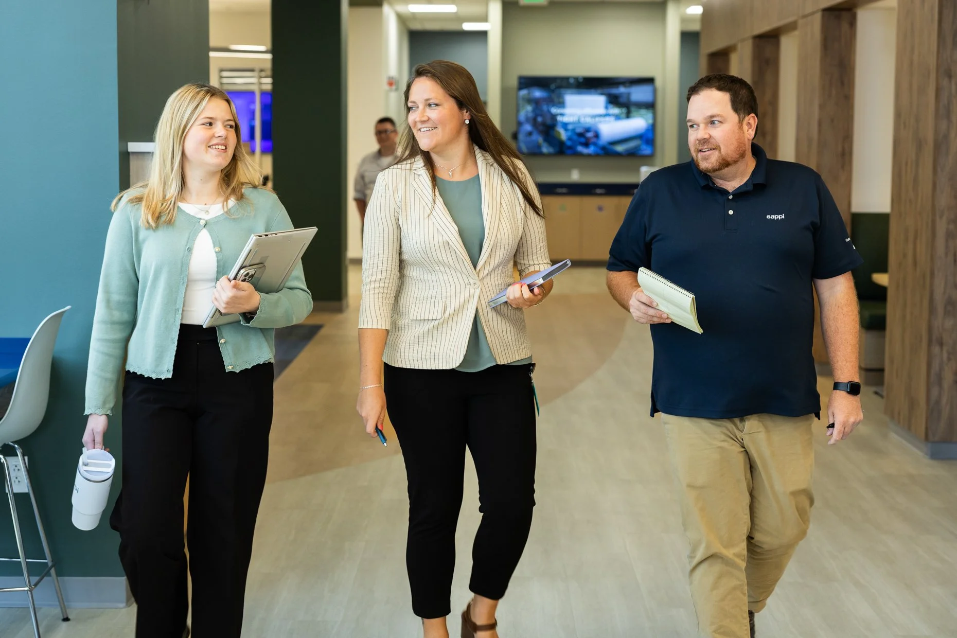 Three people, two women and one man, walking and talking in a modern indoor office or lobby environment, holding notebooks and a water bottle.