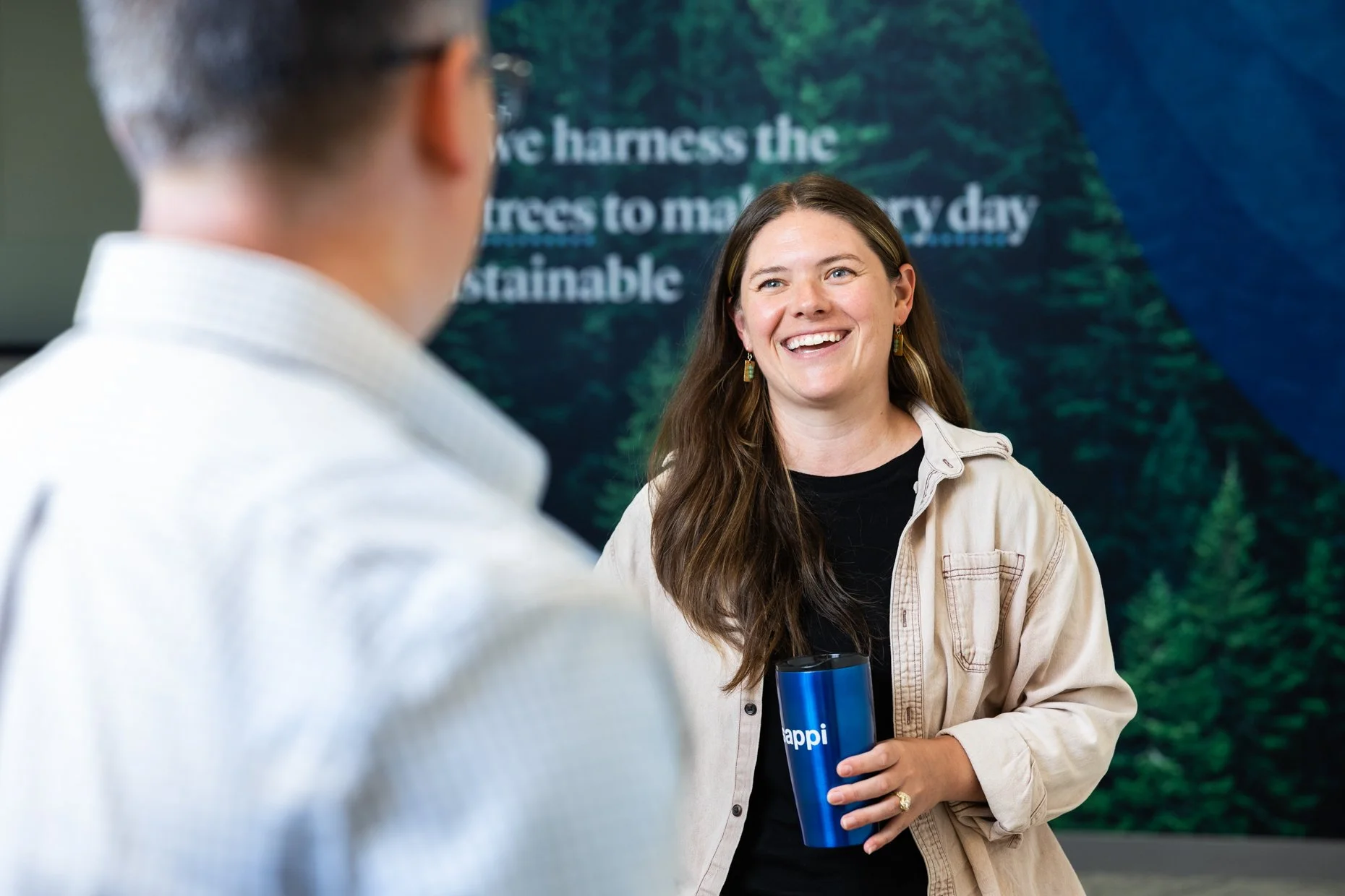 Smiling woman with long brown hair wearing a beige jacket holding a blue tumbler and talking to a man with glasses, in front of a backdrop with green trees and blue sky.