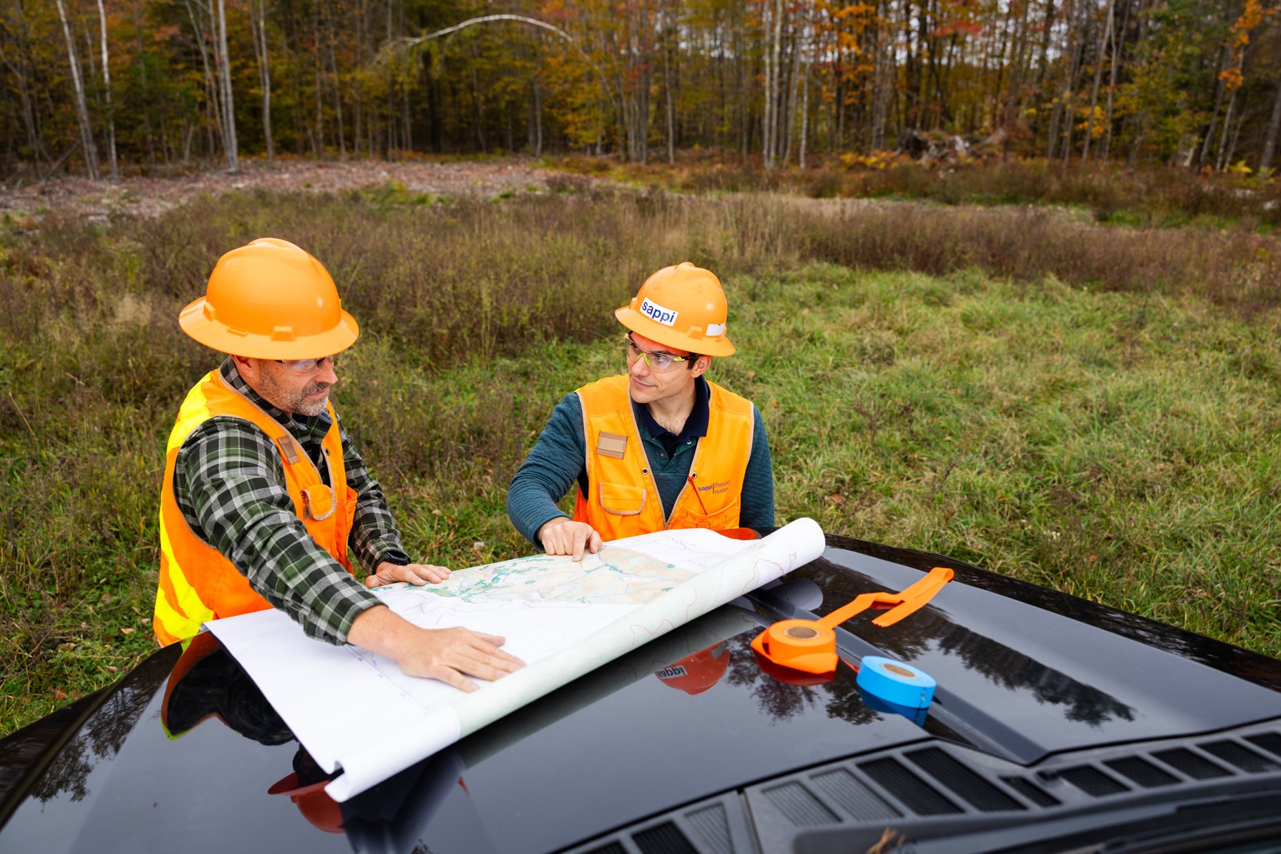Two construction workers in orange safety helmets and vests studying a large map on a black car hood in an outdoor setting with trees and autumn foliage in the background.