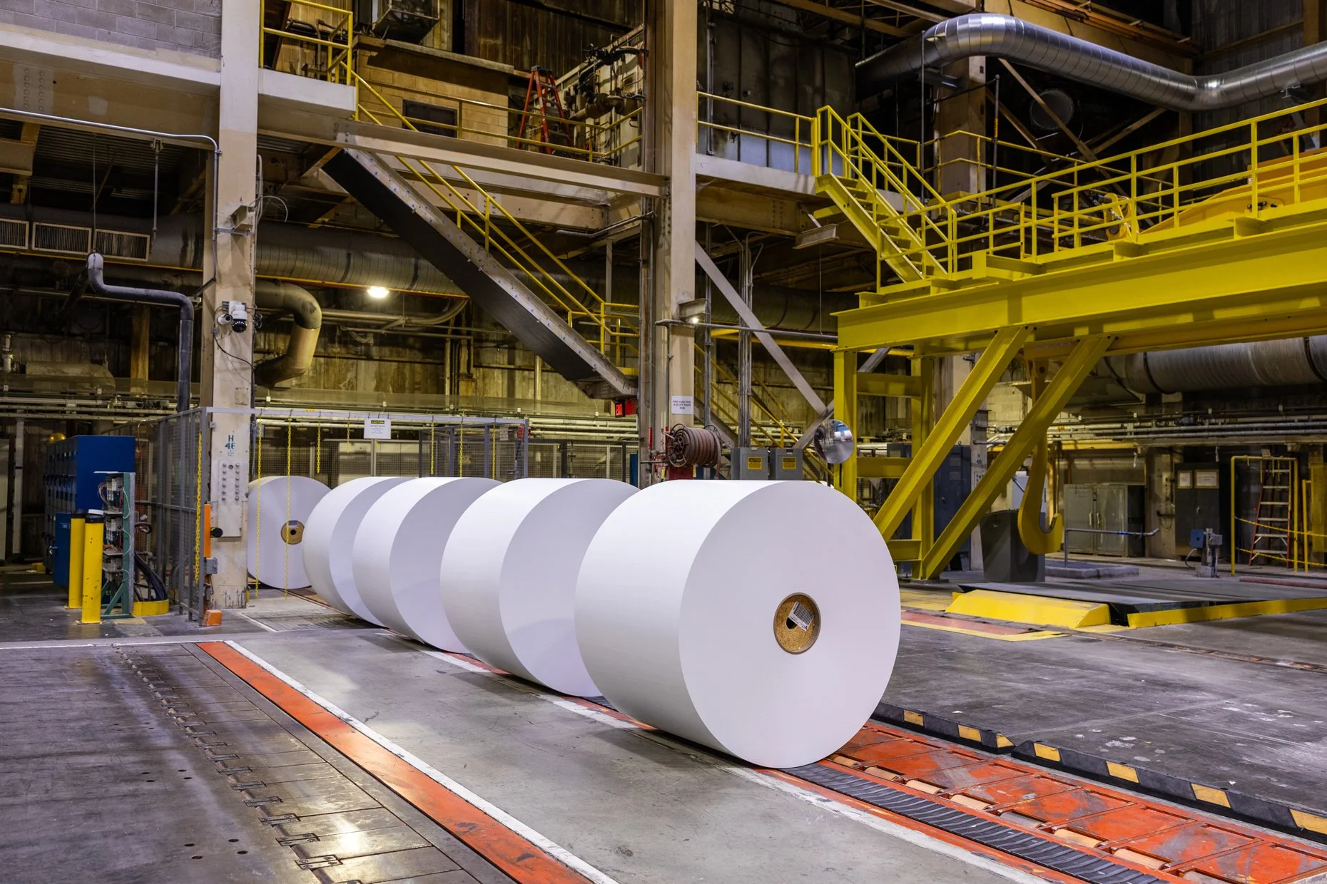 Industrial facility with large rolls of white paper and yellow machinery, including stairs and pipes, in a factory setting.