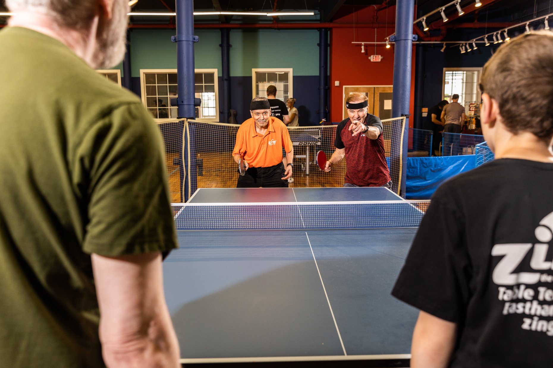 Two men with headbands playing ping pong at an indoor table, with two other people watching.