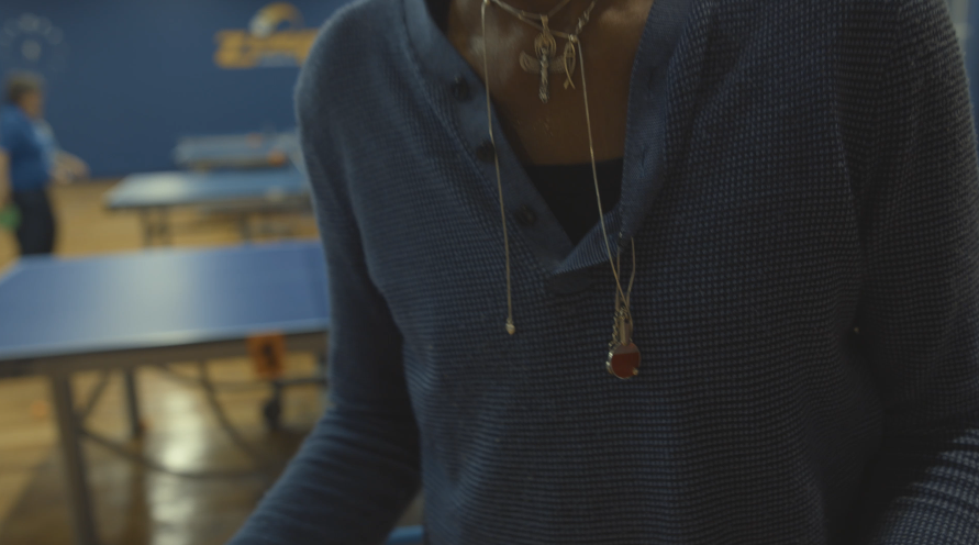 Close-up of a person wearing a dark blue textured long-sleeve shirt and a gold necklace with pendants, in a room with ping pong tables.