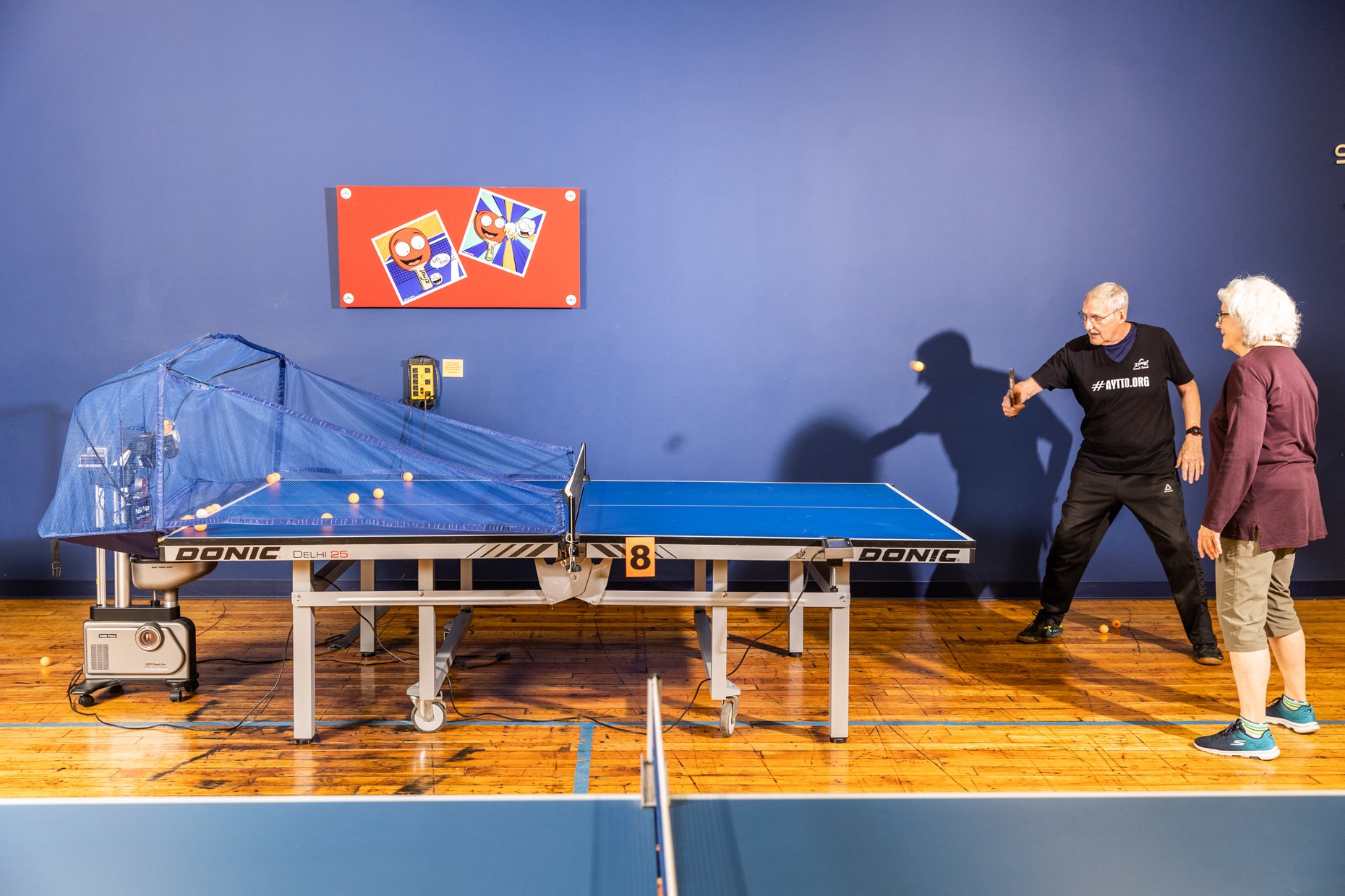 Two older adults playing ping pong at an indoor recreational facility. The man is hitting a ball while the woman watches. There is a blue wall with a colorful cartoon image of a smiling character, a ping pong table, and some balls on and around the t