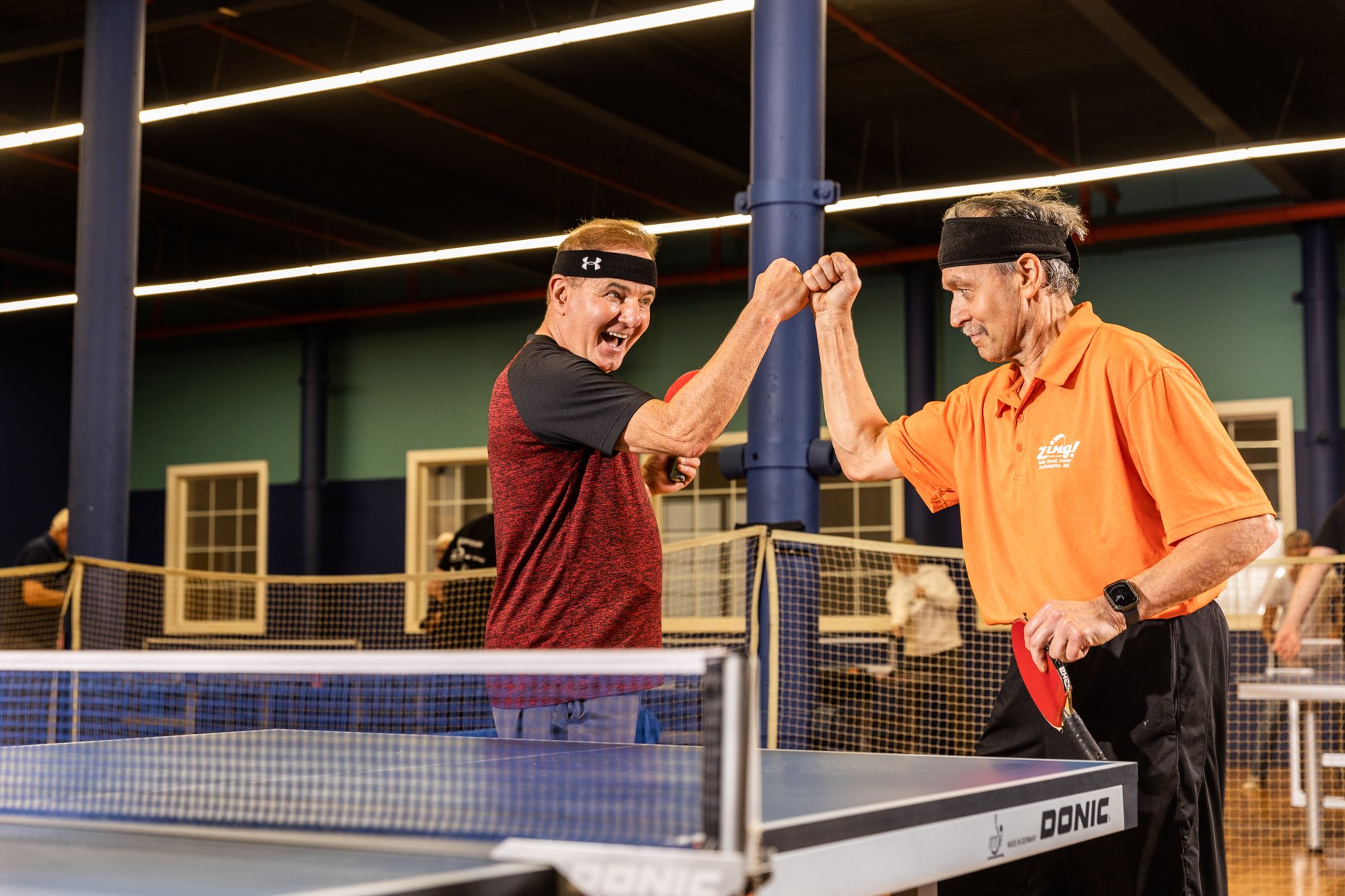 Two men with headbands are playing table tennis in a gymnasium. They are bumping fists and smiling at each other, celebrating a point. The environment is indoors with bright lighting.