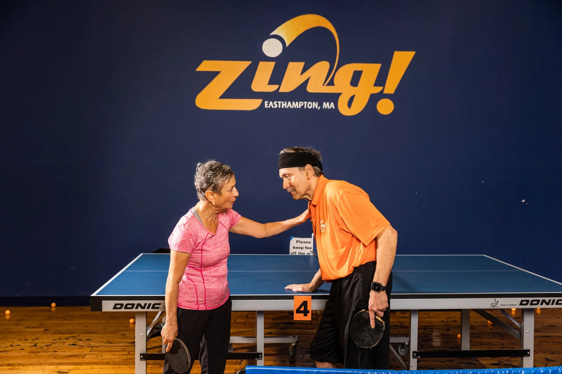 An older woman and a middle-aged man are playing table tennis in a gym or recreation center. The woman is wearing a pink athletic shirt, and the man is wearing an orange polo shirt and a black headband. They are facing each other near a ping pong tab