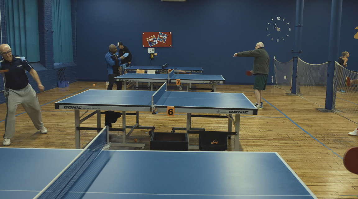 Indoor table tennis facility with several tables, players practicing, and spectators. Blue walls, a wall clock, and a colorful artwork on the wall.