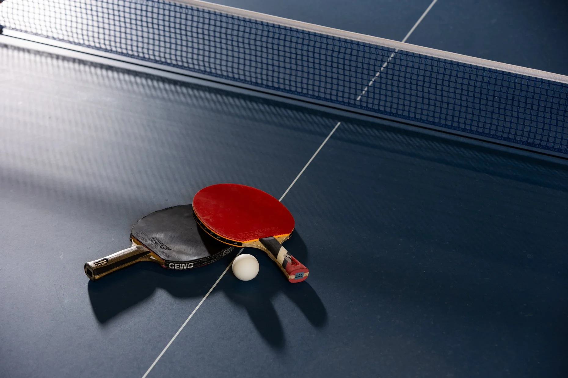 Table tennis equipment including two paddles, a white ball, and a net on a blue table.