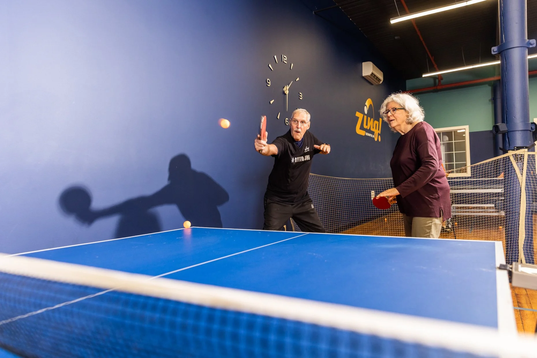 An older man playing ping pong with a woman in an indoor table tennis facility, with a blue wall and clock in the background.