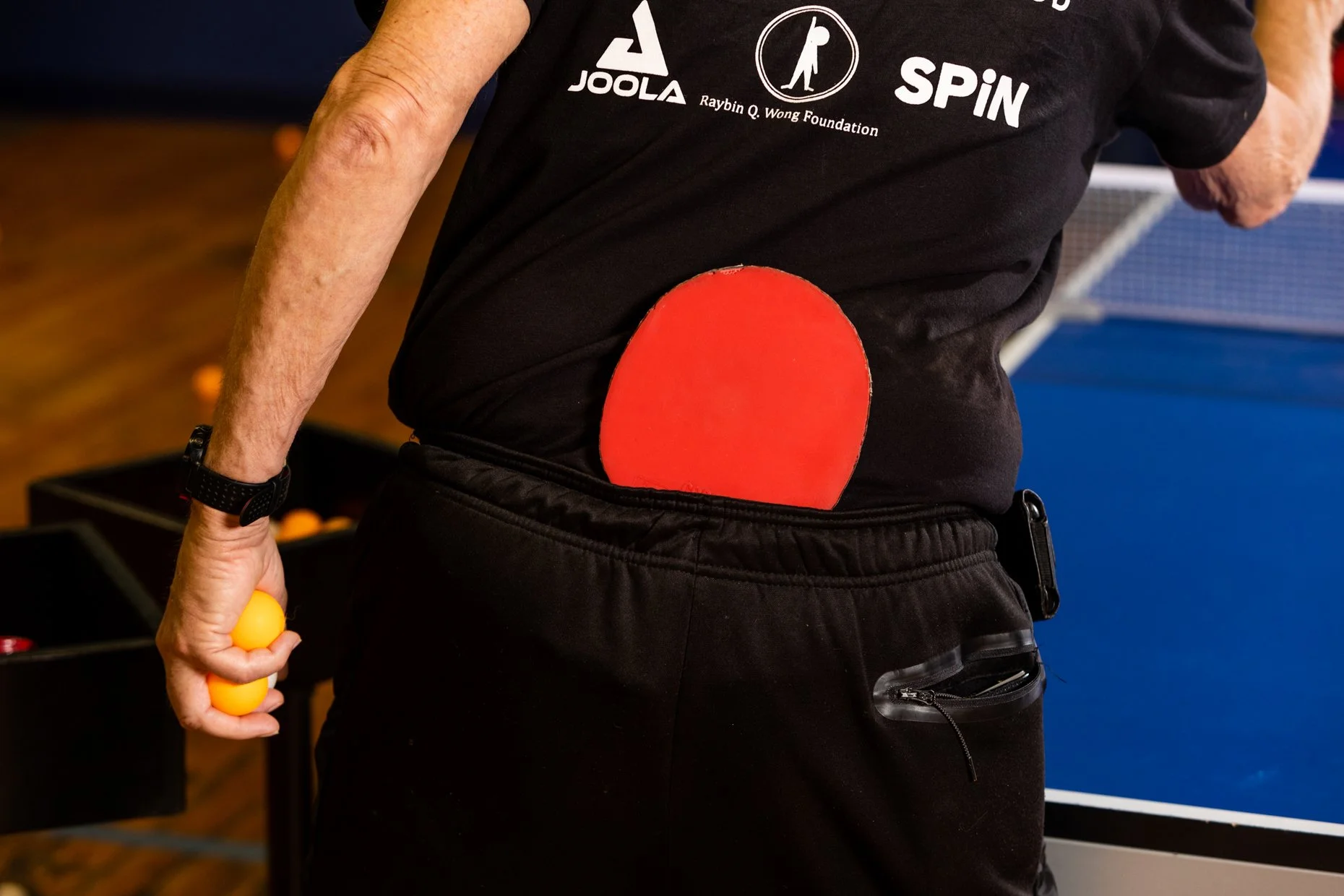 A person playing ping pong holding a yellow paddle with orange balls in the other hand. The person is wearing a black T-shirt with logos and an illustration of a person holding a racket, and black pants with a pocket. There is a red ping pong paddle 