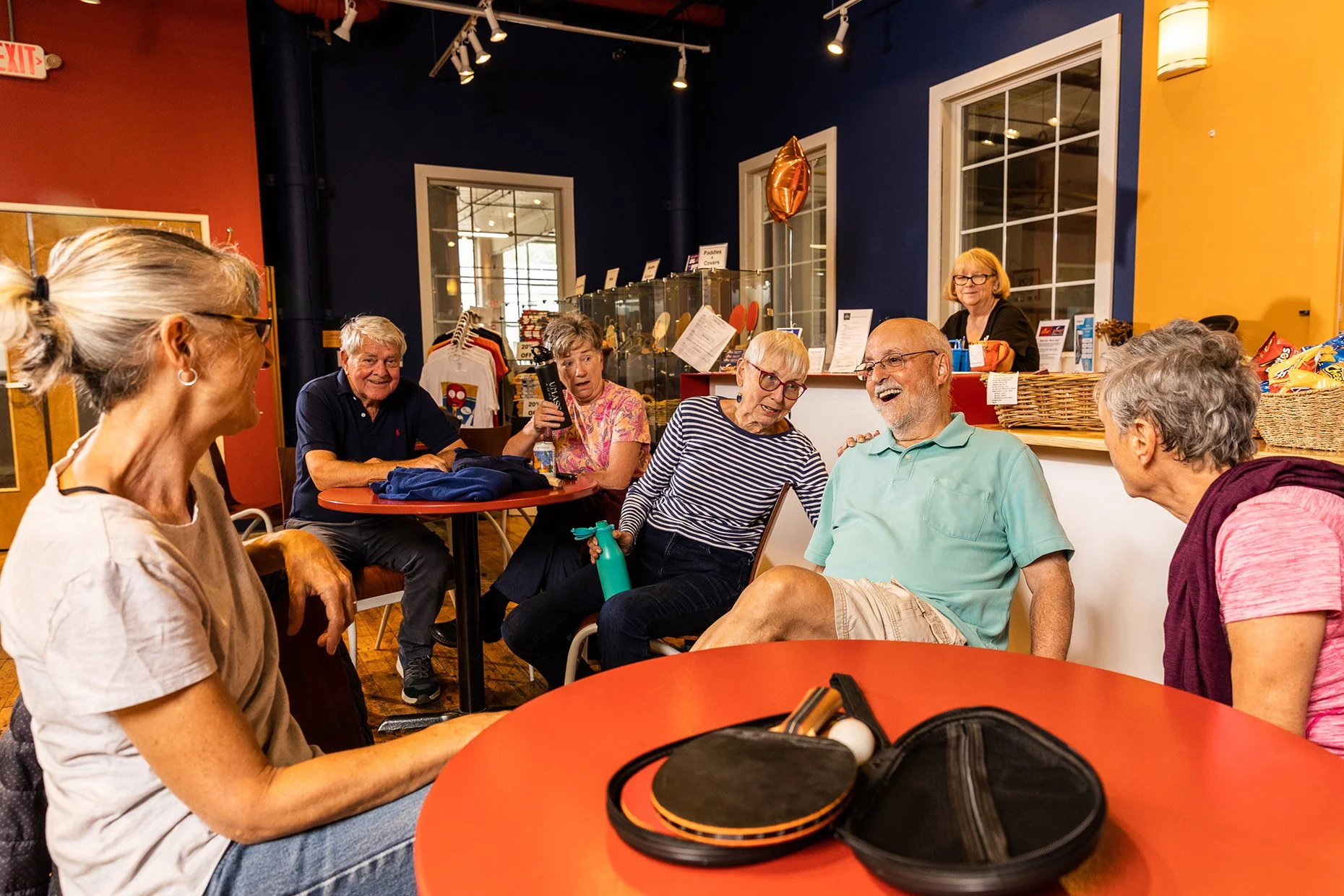 A group of elderly people sitting around tables inside a cafe or community center, engaged in conversation and laughter, with a barista behind the counter in the background.