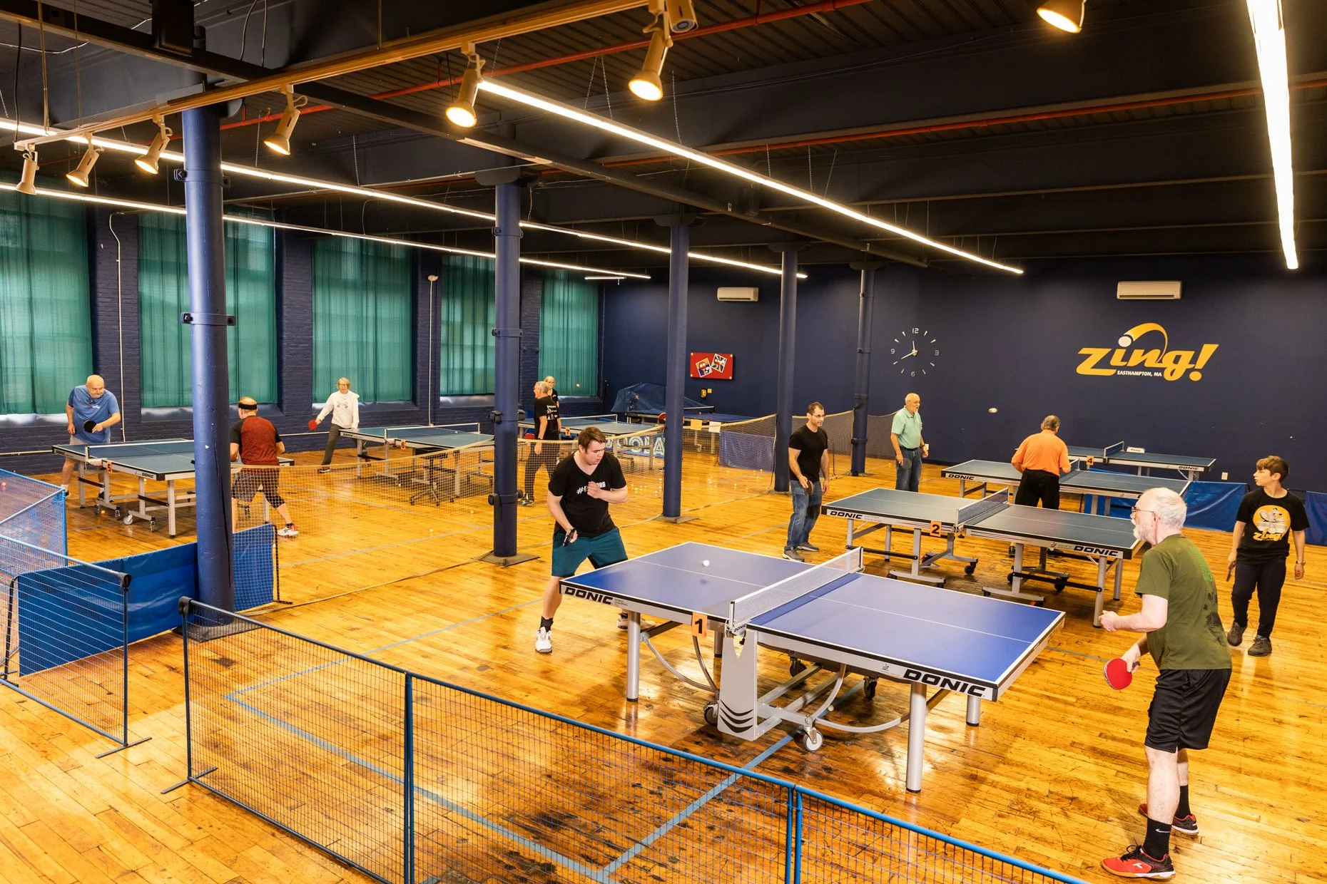 Indoor ping pong facility with multiple table tennis tables in use. Several people are playing or standing around, some with paddles. The room has wooden floors, blue walls with large green curtains, and a wall clock. Bright overhead lighting and a l