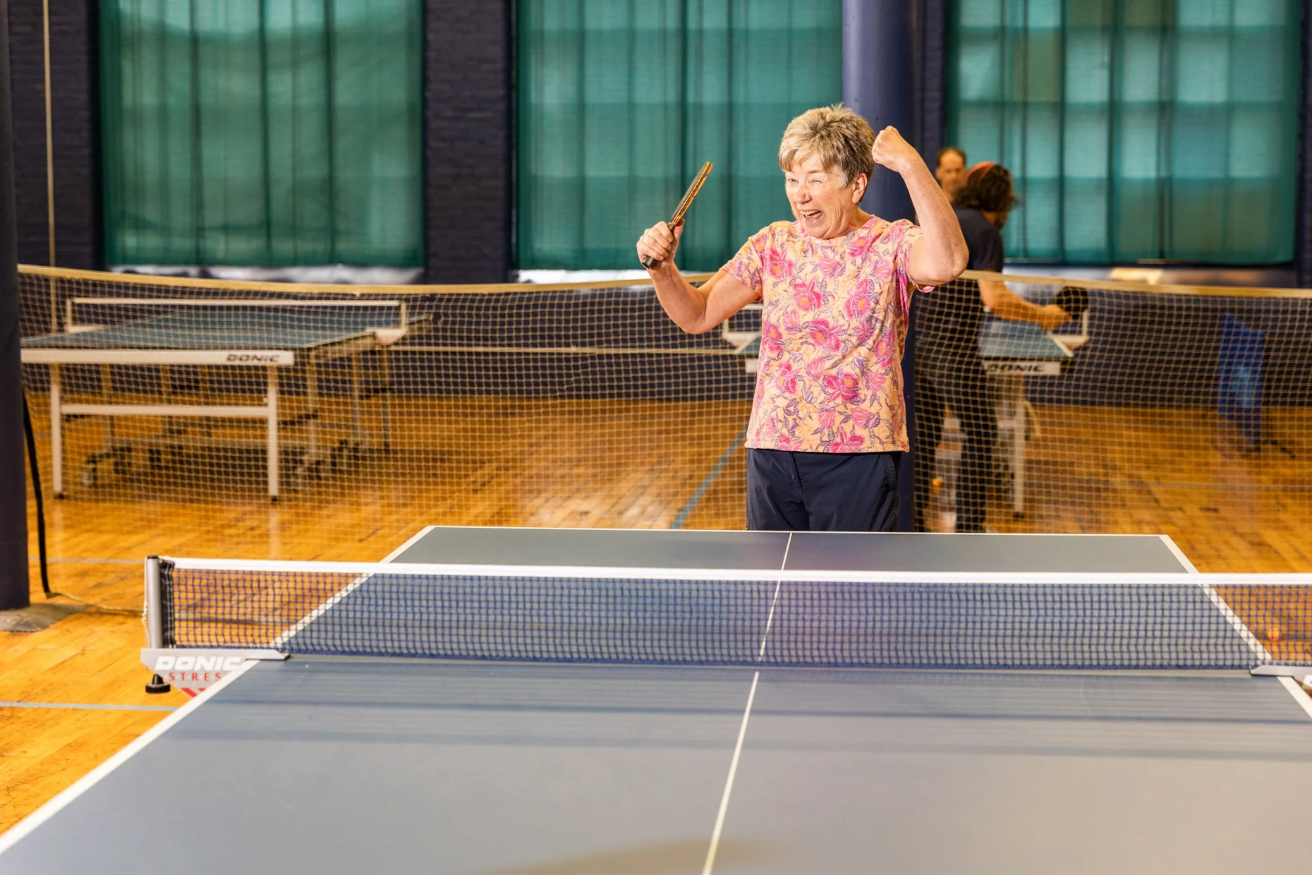 An elderly woman celebrating victory after winning a table tennis match, holding a paddle and raising her arm in a gymnasium with multiple ping pong tables and green curtains.