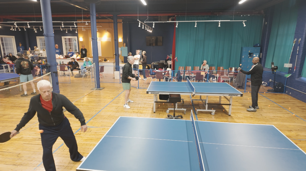 People playing ping pong in a recreational room, with others sitting and socializing in the background.