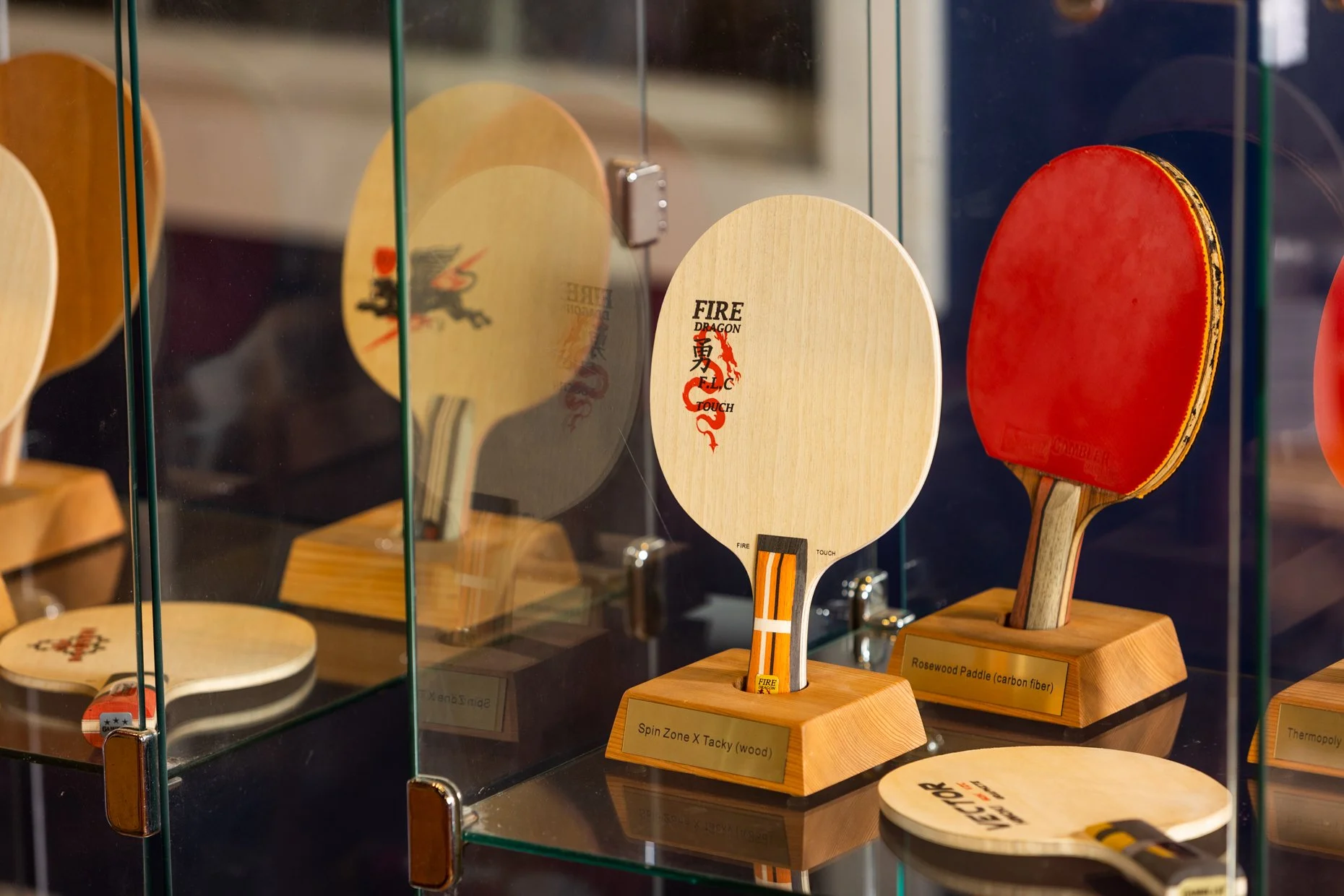 Display of ping pong paddles in a glass case, with labels indicating their materials and brands.