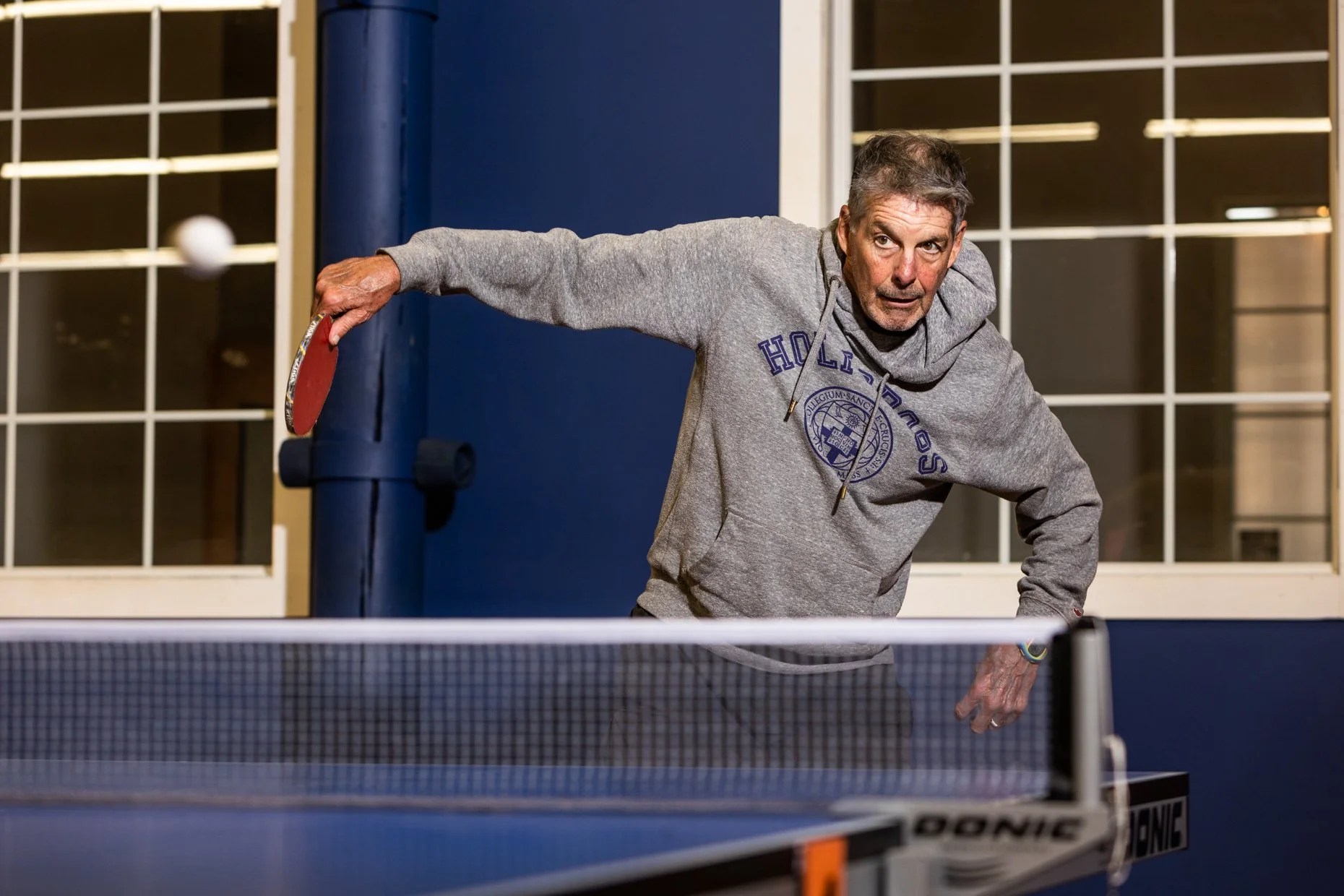 An older man playing table tennis indoors, wearing a gray hoodie with purple lettering, arm extended with a paddle, ready to hit the ball.