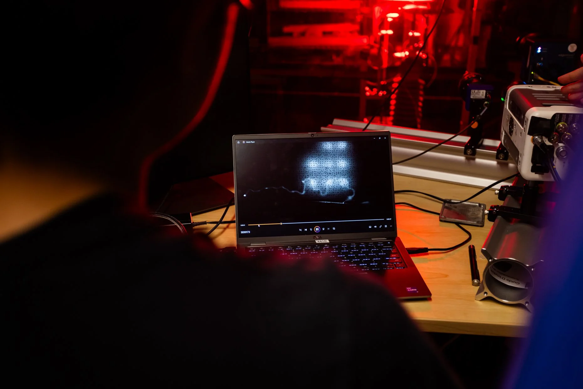 A laptop on a wooden table displaying a waveform on a media player. To the right, equipment including a white electronic device with cables and a small camera on a tripod are visible. The background shows a red-lit environment with visible electronic
