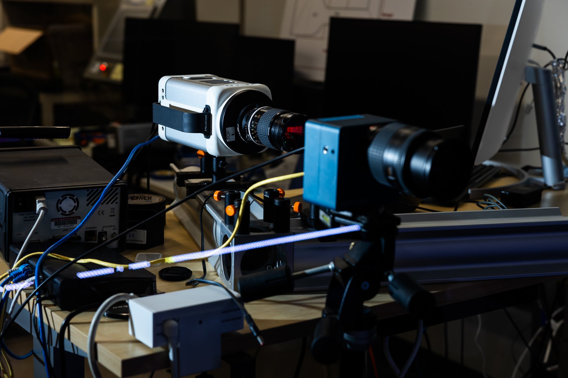 Laboratory setup with camera mounted on a motorized track, connected to various electronic devices and wires on a wooden table, in a dimly lit room with multiple monitors and equipment in the background.