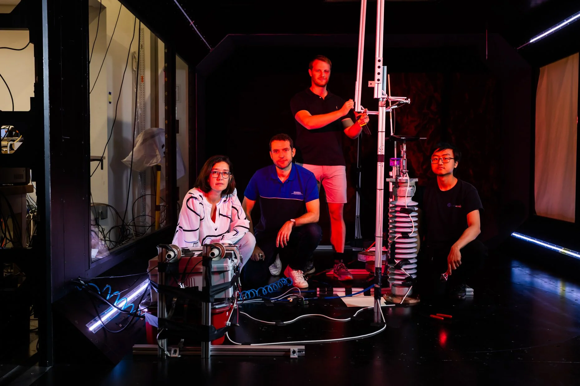 Four people in a dark room with scientific equipment. Two are sitting, one is kneeling, and one is standing. They are surrounded by wires and devices, with some illuminated pink and blue lights.