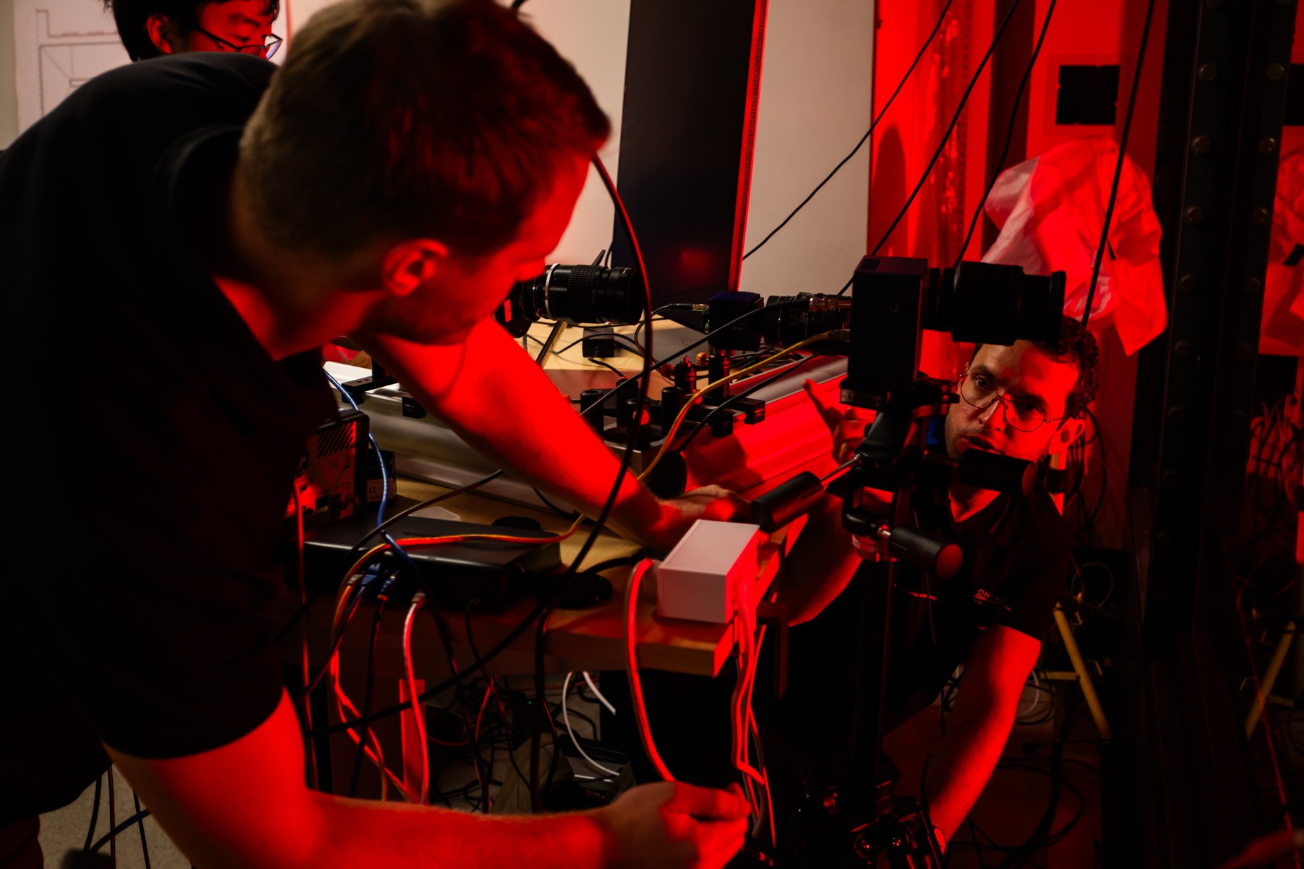 Scientists working with a complex laser setup in a lab, illuminated by red light, with equipment, wires, and devices around them.