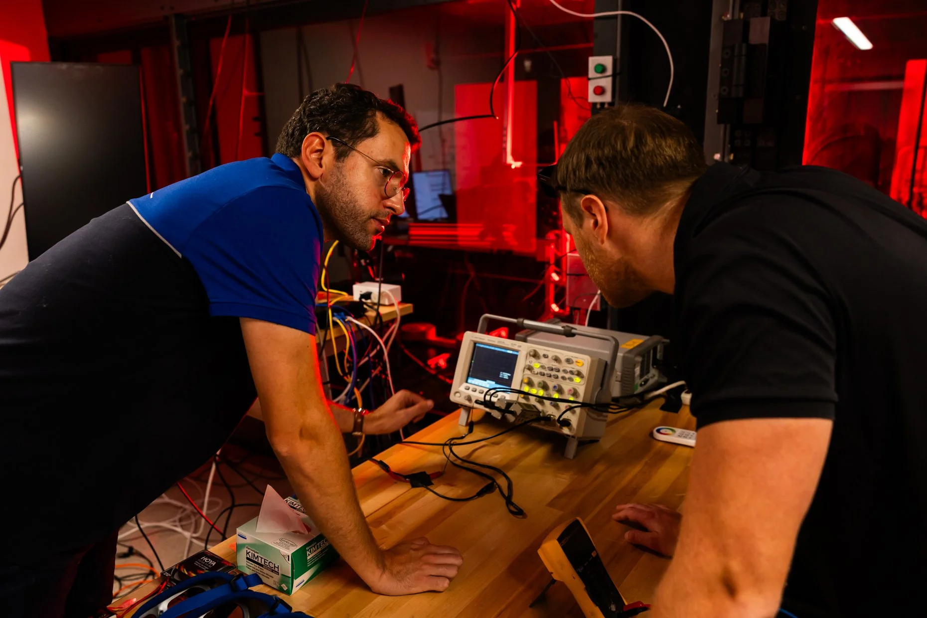 Two men working with electronic equipment in a lab, bent over a wooden table with wires and a testing device, illuminated by red lighting.