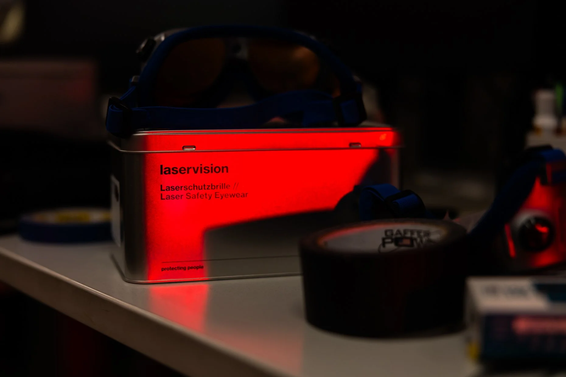 A metal red tin box labeled 'laservision' containing laser safety eyewear, placed on a surface with other items in a dimly lit environment.