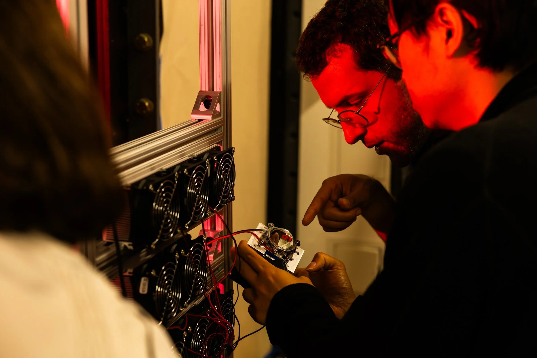 Two men working on a cryptocurrency mining rig with multiple cooling fans and red wiring.