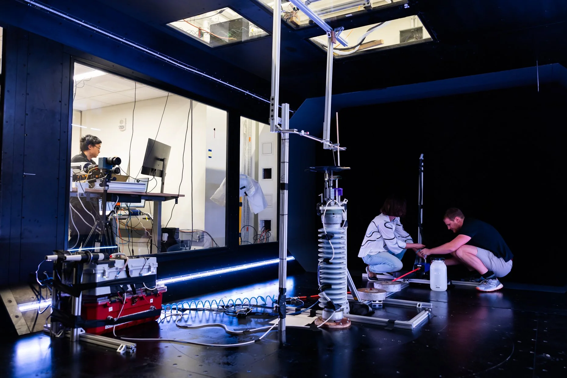 Two people are working on a scientific experiment on a black stage while another person is working at a computer from a glass-walled room. The scene includes various scientific equipment, metal structures, and wires, with some people kneeling and oth