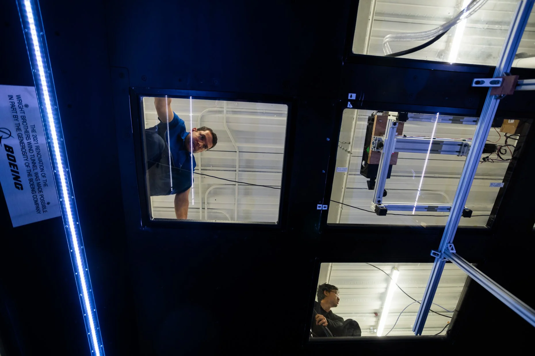 People working inside a controlled environment chamber or laboratory with glass windows, metal frames, and lighting, observed from below.