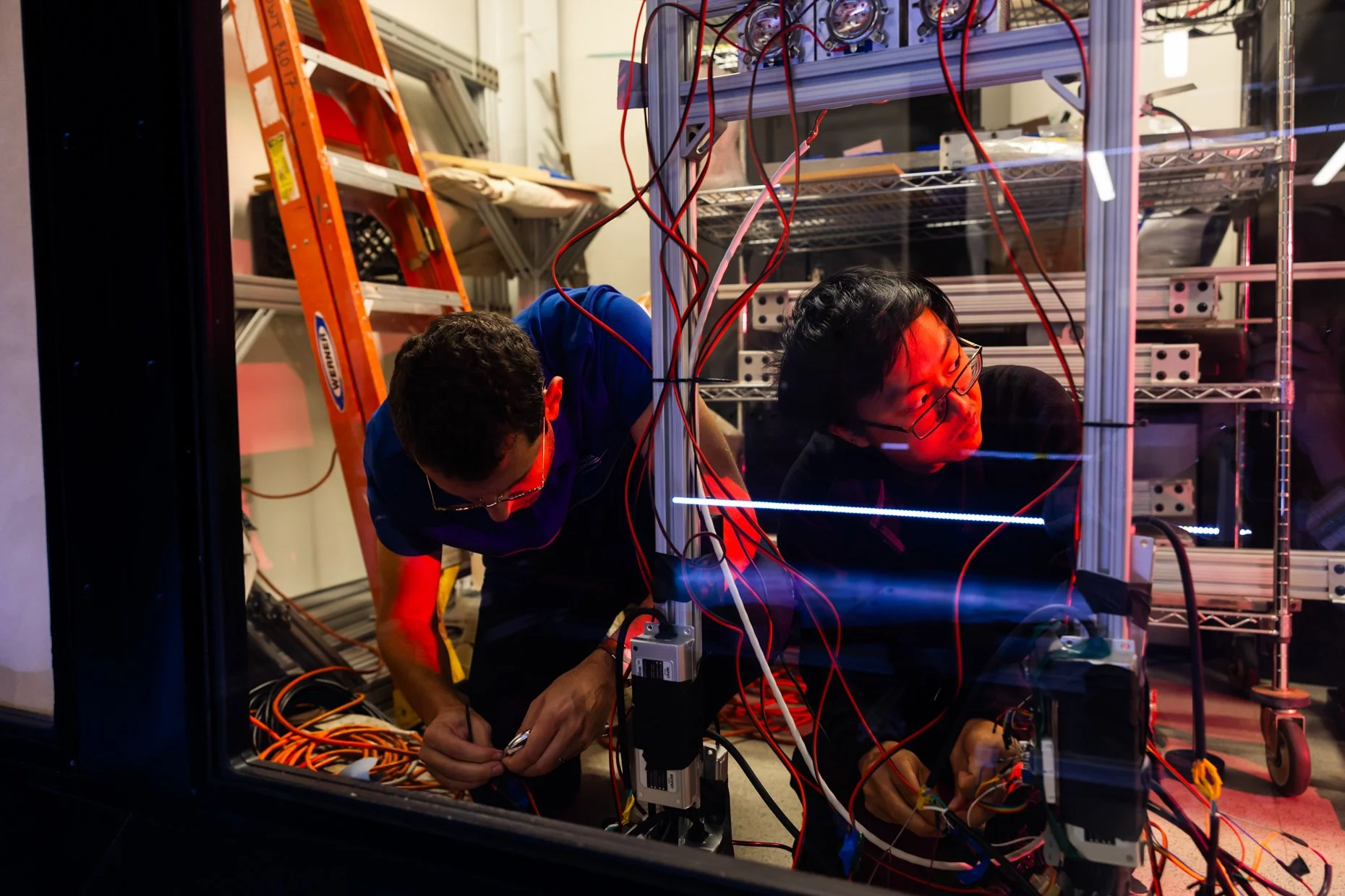 Two researchers working inside a lab setup with metal racks, wiring, and electronic equipment, focused on a project involving red and blue LEDs.