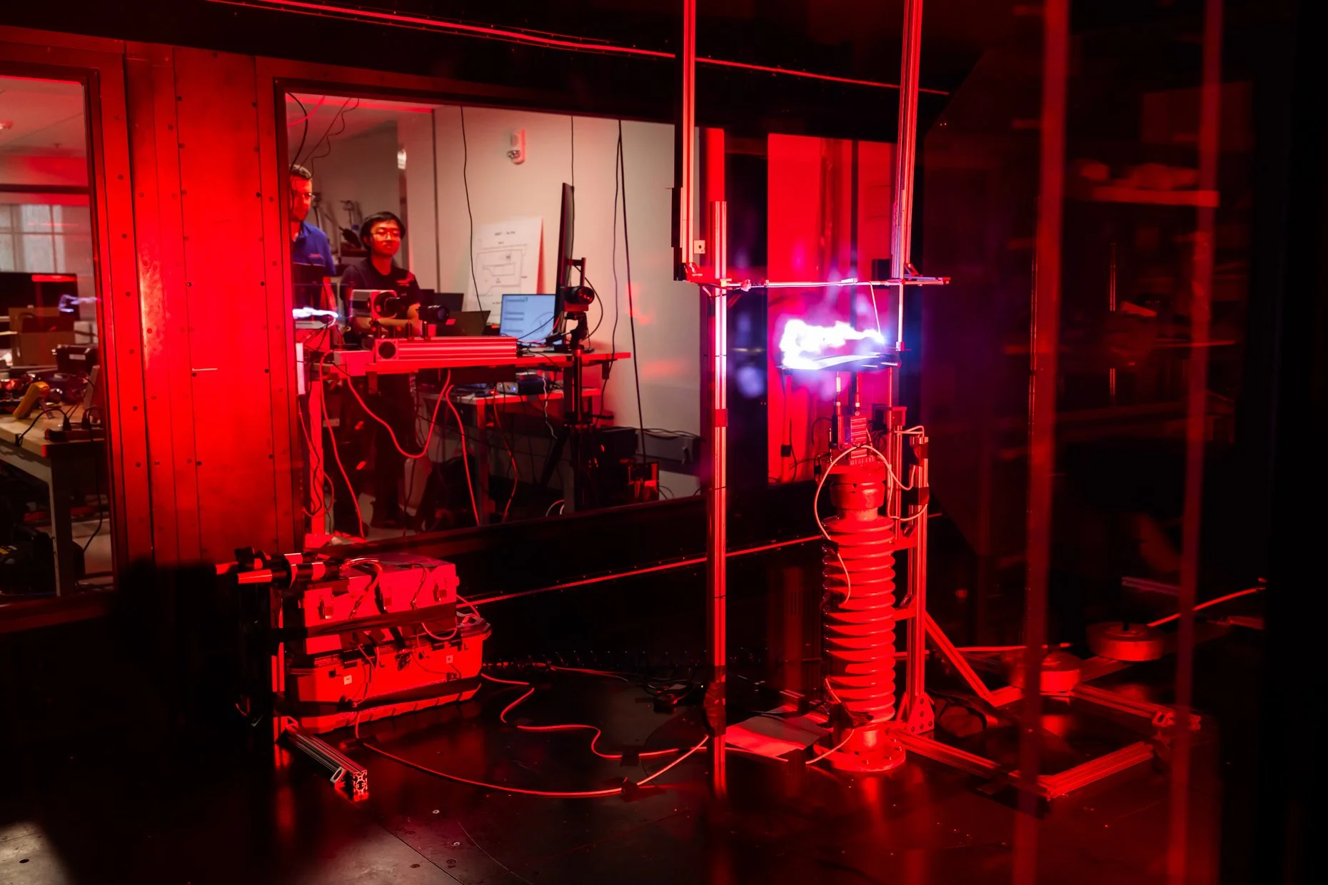 Laboratory setting with two scientists working on a setup involving red laser equipment, mirrors, and optical components, inside a secured glass enclosure.