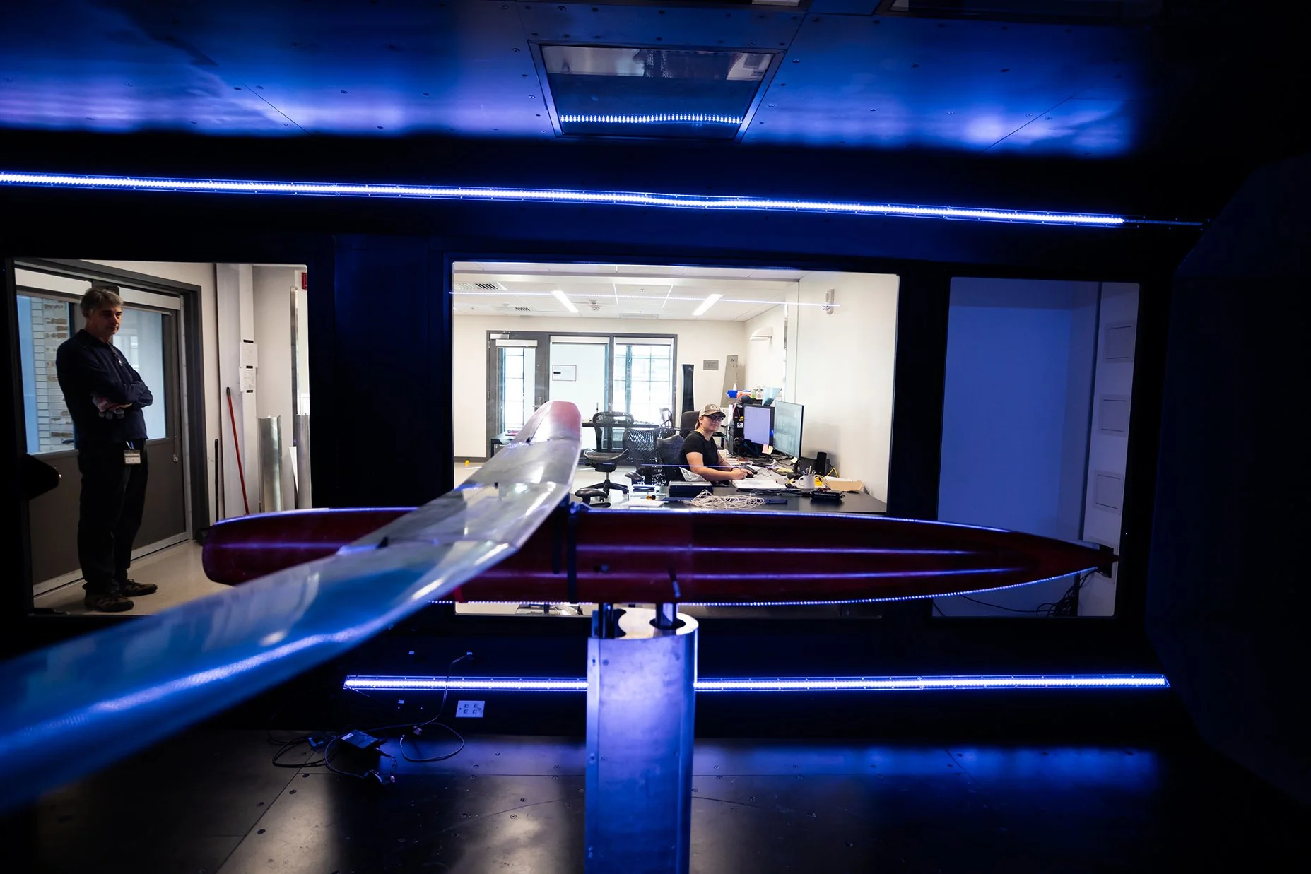 Interior view of a room through a window showcasing a woman working at a desk with dual monitors, a man standing by the door, and various electronic equipment and cables. The scene features blue and purple lighting accents.