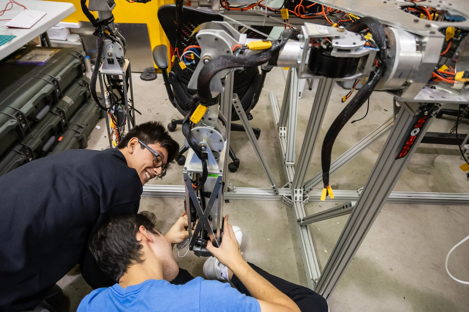 A group of four young men working on assembling or repairing a robot in a workshop. They are focused on the robot's frame, which is made of metal and surrounded by wires and electronics. The workspace has tools and equipment, and the men are using th