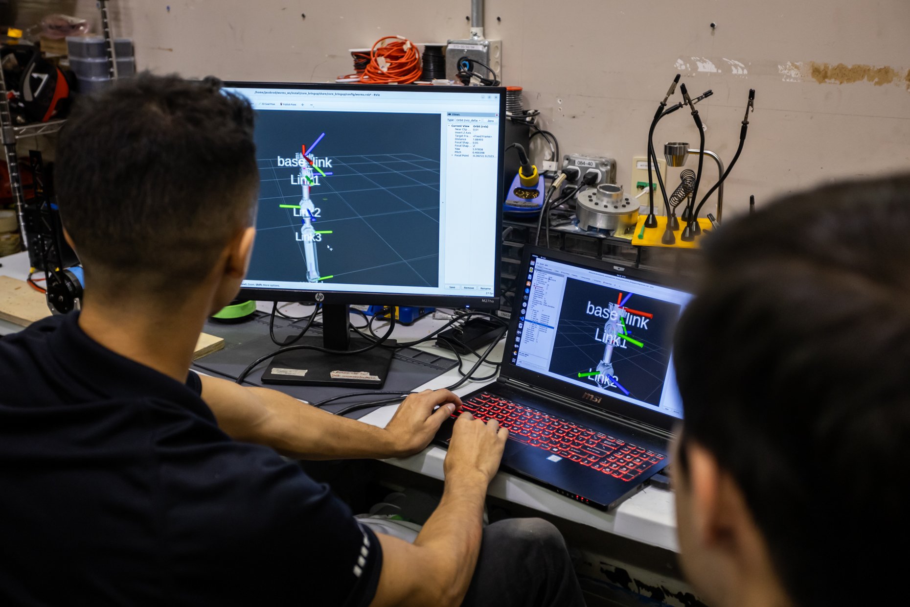 People working on computers with 3D modeling and simulation software related to robotics or engineering, with equipment and wires in the background.