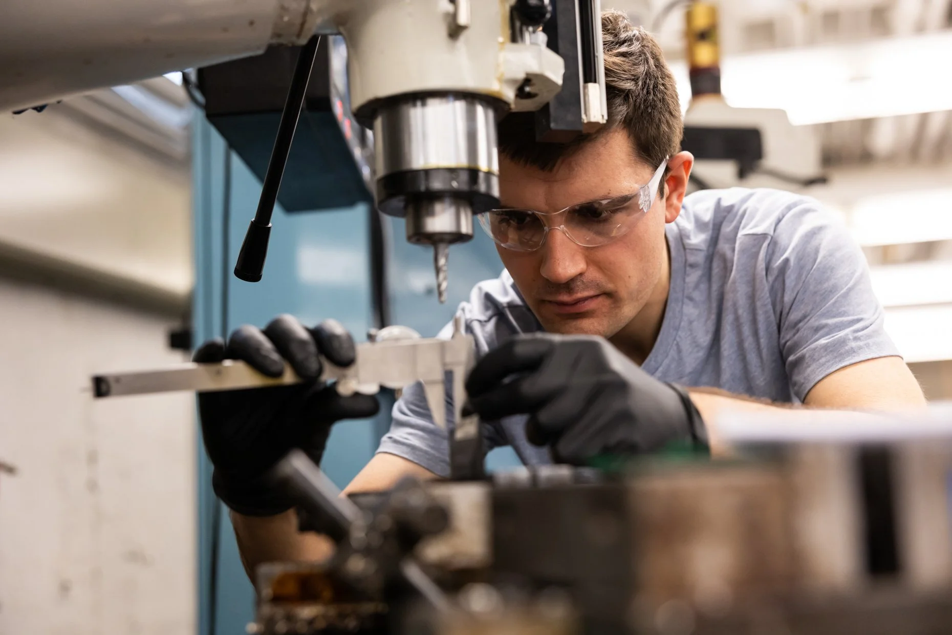 A man wearing safety glasses and gloves working with a drill press in a workshop or lab environment.