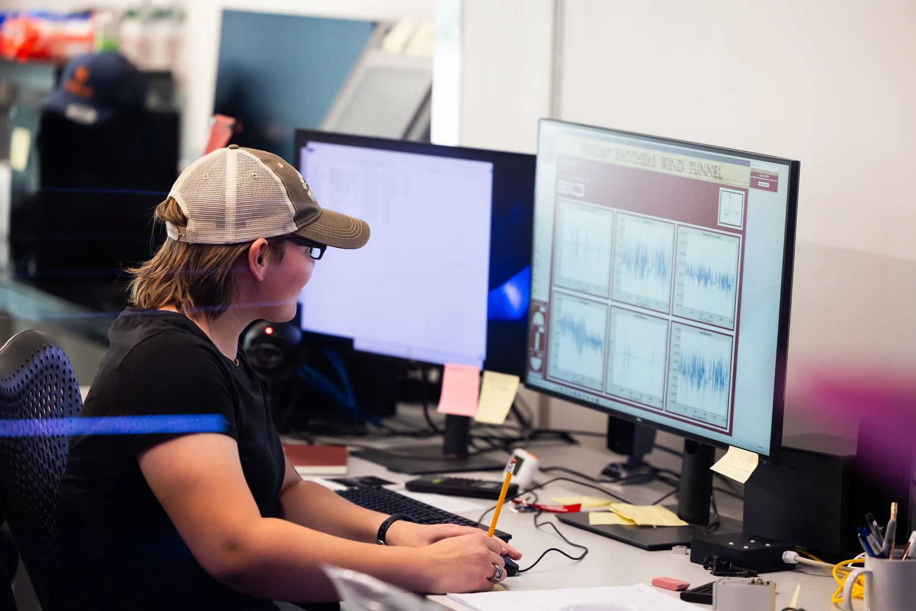 A person wearing a beige mesh cap and black shirt is working at a desk with two large computer monitors, analyzing data visualizations and charts on the screens.